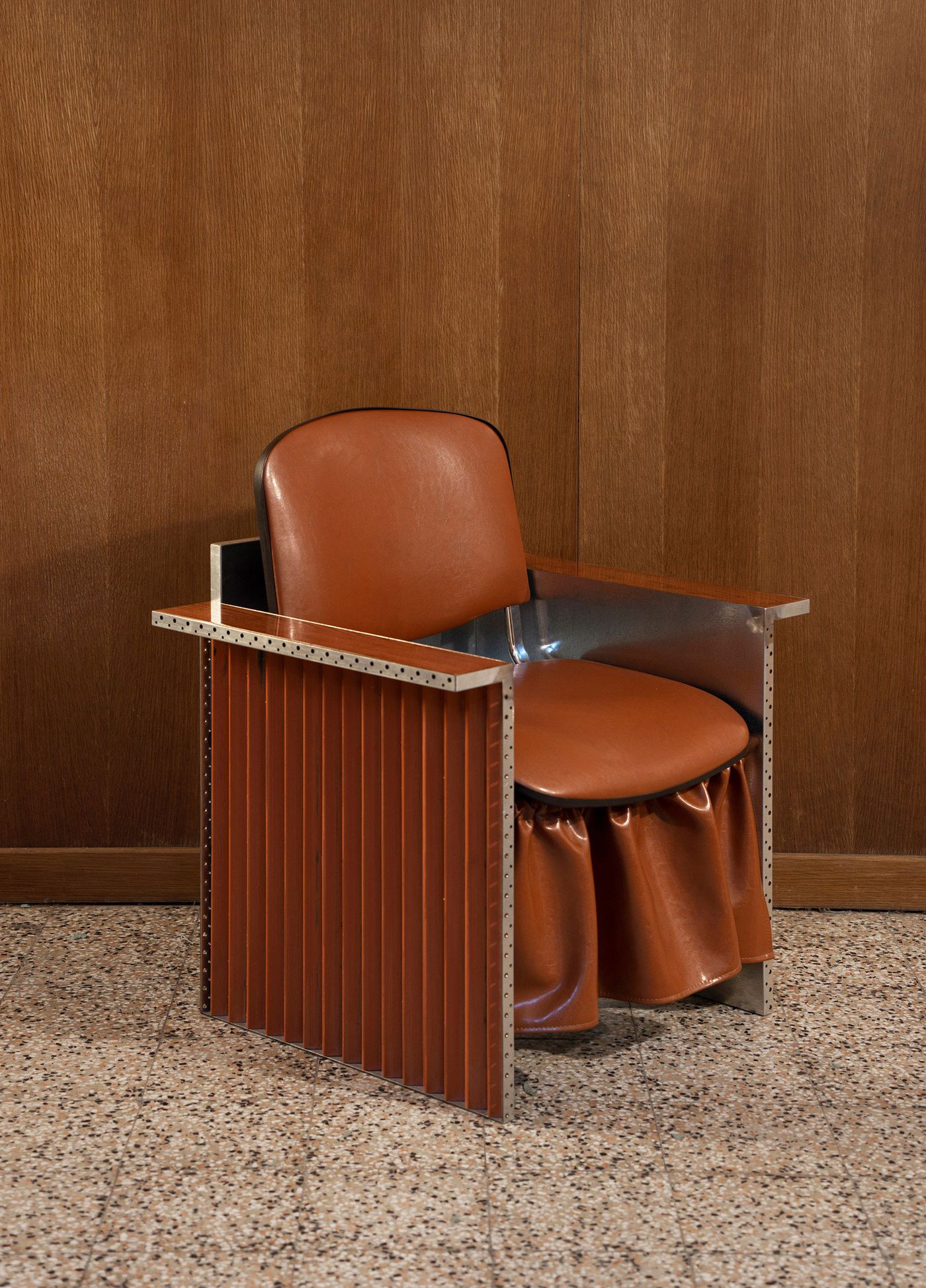 A close-up of a single leather and metal armchair with a gathered leather skirt detail, set against a wood-paneled wall and terrazzo floor.