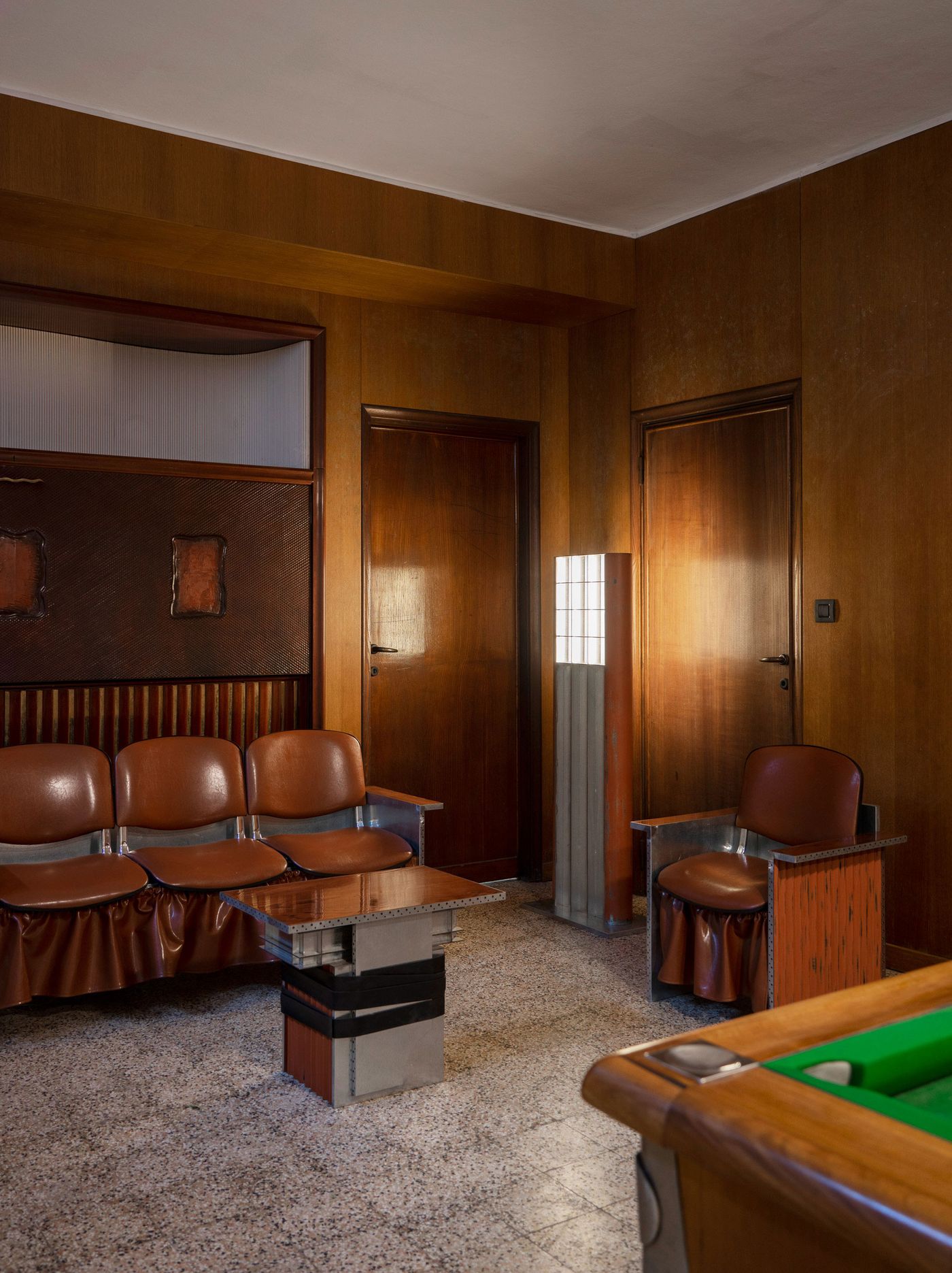 A vintage-style waiting room with warm wood-paneled walls, leather and metal chairs, a metallic table, and a tall grid-like floor lamp.