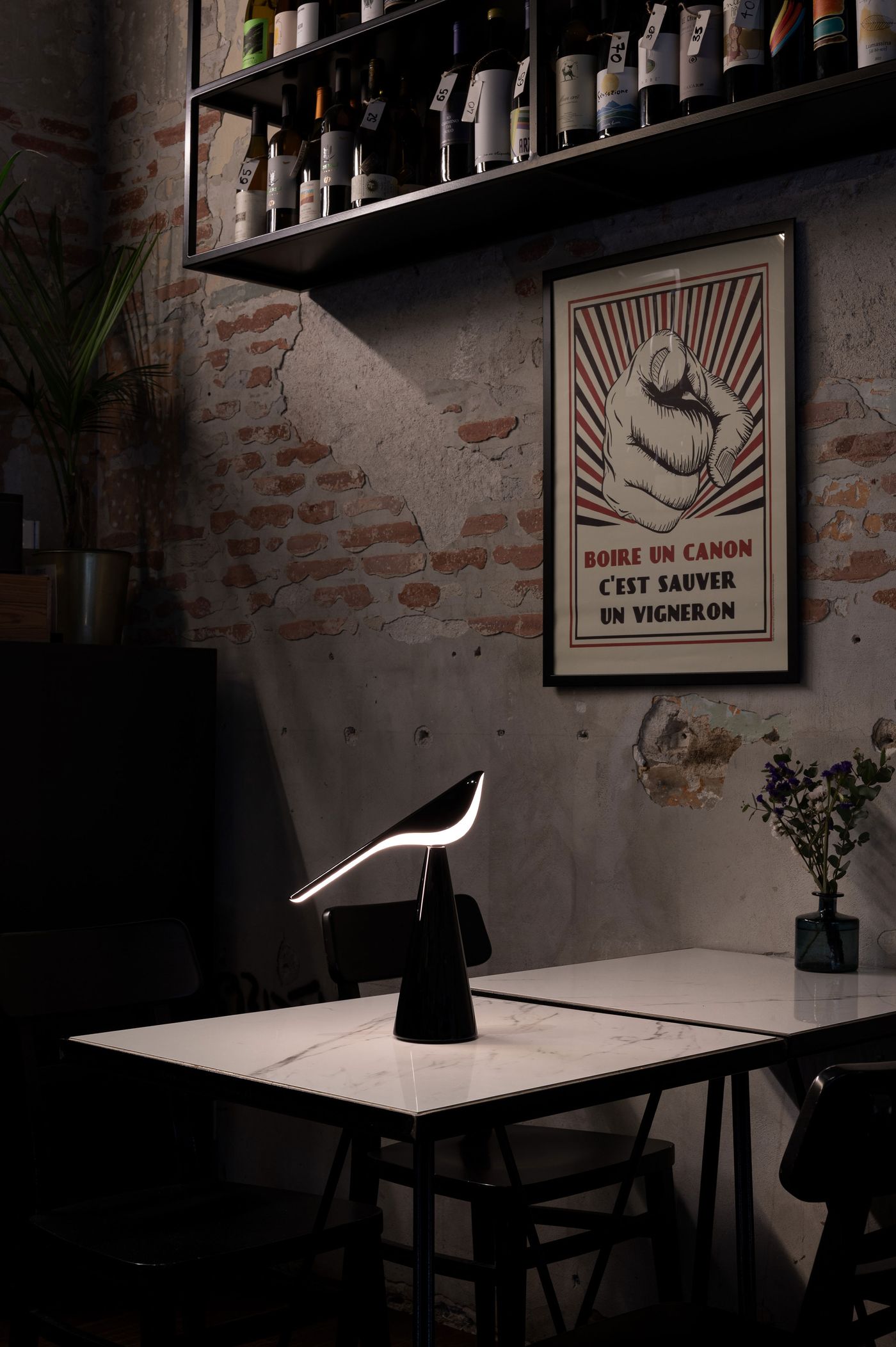 A sleek black table lamp in the sculptural shape of a perched bird, designed by Nao Tamura for Established & Sons, sits on a white marble tabletop in a moody, industrial-style bar with exposed brick walls, a framed vintage French poster, and bottles of wine overhead. A sleek black table lamp in the sculptural shape of a perched bird, designed by Nao Tamura for Established & Sons, sits on a white marble tabletop in a moody, industrial-style bar with exposed brick walls, a framed vintage French poster, and bottles of wine overhead.