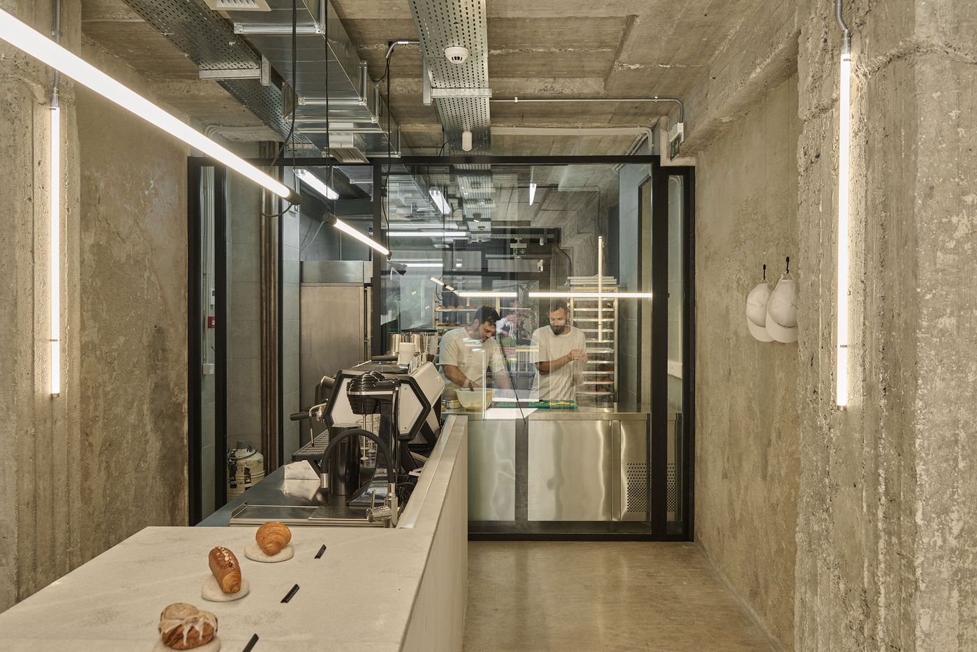 The industrial-minimalist interior of Temps Perdu bakery features rough concrete walls, exposed ceiling services and linear fluorescent lighting. A pale stone display counter showcases a few artisanal pastries, while a glass partition reveals two bakers working in a stainless-steel preparation area, highlighting the contrast between raw architectural textures and precise pastry-making.