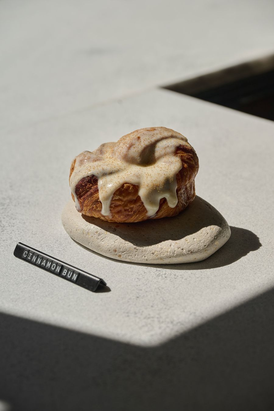 A close-up of a cinnamon bun at Temps Perdu bakery in Athens, its golden laminated pastry generously coated in thick, softly dripping white icing and dusted with cinnamon. Presented on a small, irregularly shaped stoneware plate atop a pale textured surface, the pastry is illuminated by direct sunlight that casts crisp, graphic shadows.