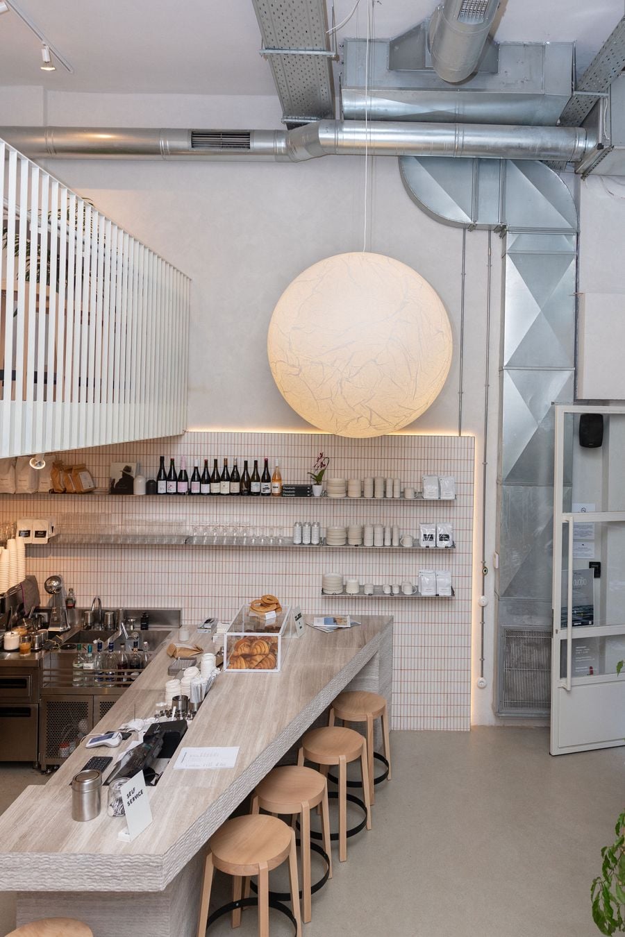 The minimalist interior of The Greek Foundation café in Thessaloniki combines a sculptural pale timber counter, matching wooden stools and white tiled walls with exposed metallic ventilation ducts. An oversized glowing spherical pendant light anchors the double-height space, lending warmth and softness to its restrained industrial aesthetic.
