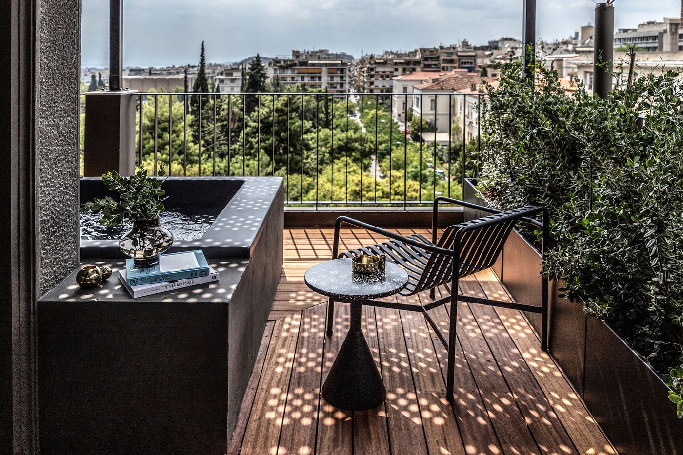 A private terrace at The Modernist hotel in Athens features a compact dark stone plunge pool, timber decking, a black metal lounge chair and a sculptural pedestal side table. Dense planting in geometric planters provides privacy, while dappled sunlight creates intricate shadows across the floor. Beyond the railing, leafy treetops give way to the city's apartment buildings.