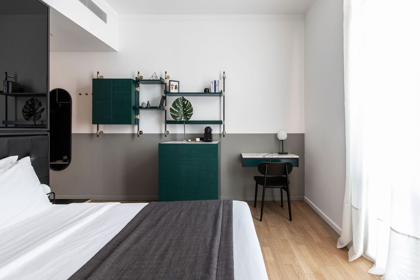 A contemporary guest room at The Modernist Thessaloniki features a restrained palette of white, charcoal grey and deep green, punctuated by brass accents and warm timber flooring. Modular wall-mounted shelving, a compact marble-topped desk, black furniture and sheer curtains create a refined, Scandinavian-inspired atmosphere.
