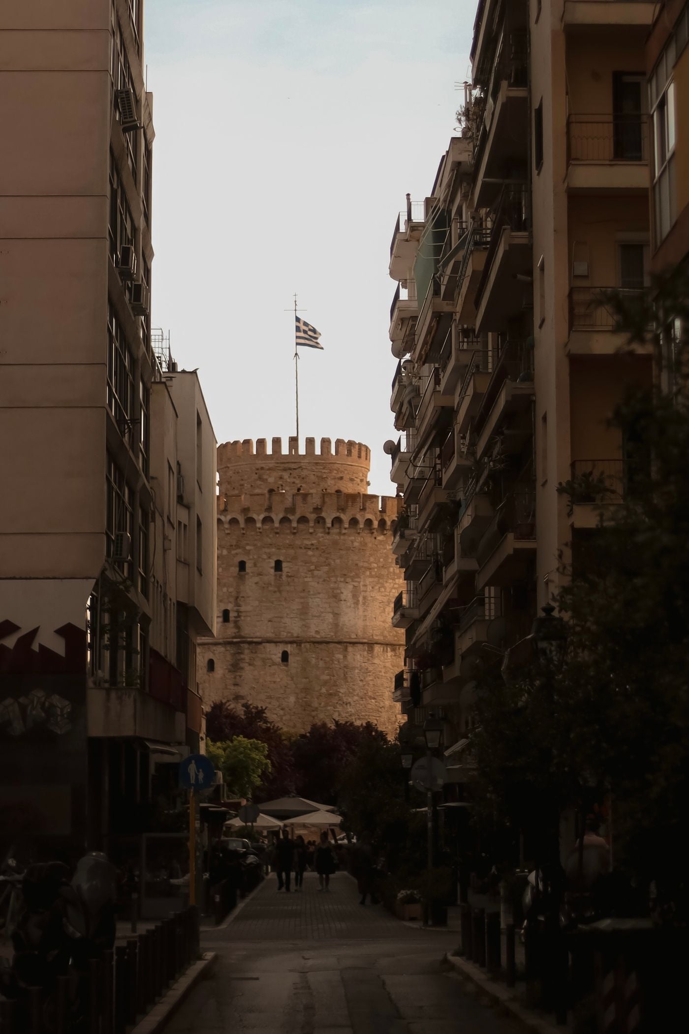 Thessaloniki’s iconic White Tower appears at the end of a narrow urban street, framed by closely packed apartment buildings and projecting balconies. Bathed in warm late-afternoon light, the medieval stone landmark rises above the shaded street, its crenellated crown and Greek flag standing out against the pale sky.