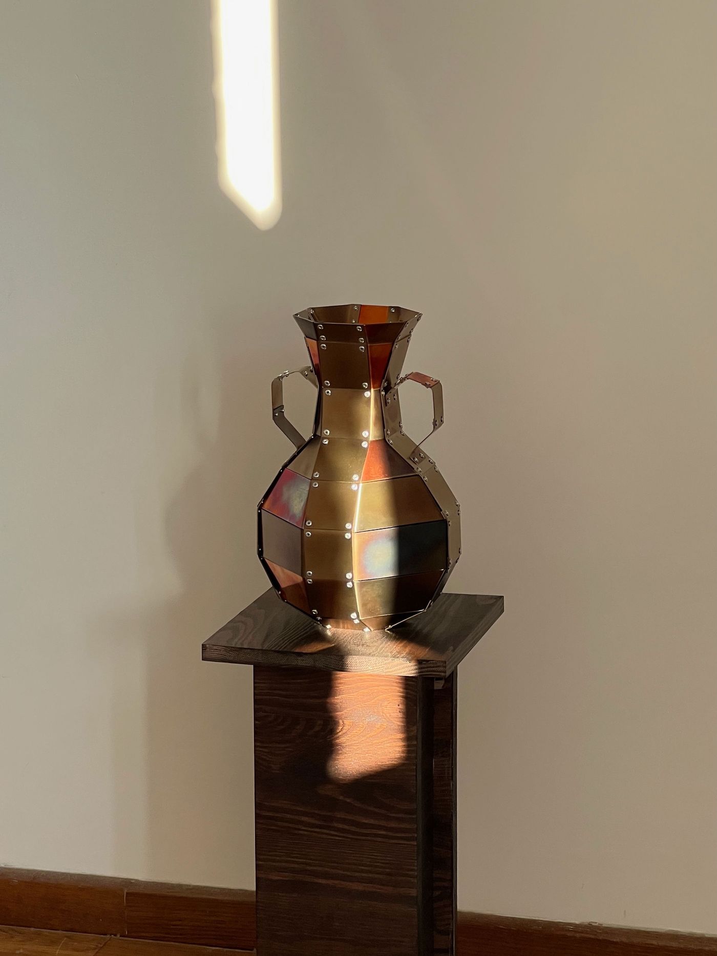 A faceted, double-handled vase crafted from riveted metal plates gleams in a beam of sunlight, casting geometric shadows on the wooden pedestal. A faceted, double-handled vase crafted from riveted metal plates gleams in a beam of sunlight, casting geometric shadows on the wooden pedestal.