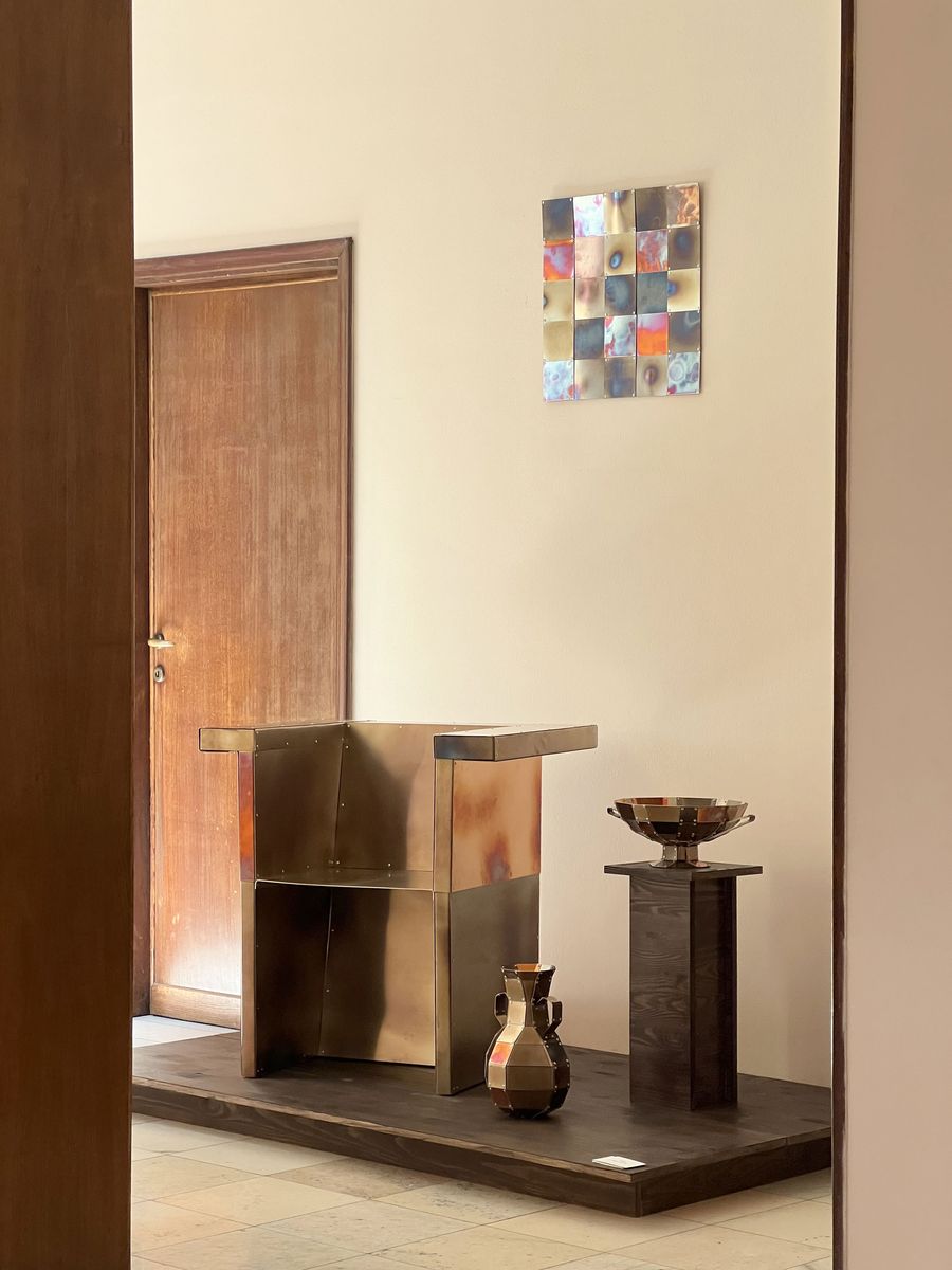 A polished metal chair and matching vessels sit atop a dark wood platform, reflecting soft warm light, beneath a pixel-like artwork of iridescent squares. A polished metal chair and matching vessels sit atop a dark wood platform, reflecting soft warm light, beneath a pixel-like artwork of iridescent squares.