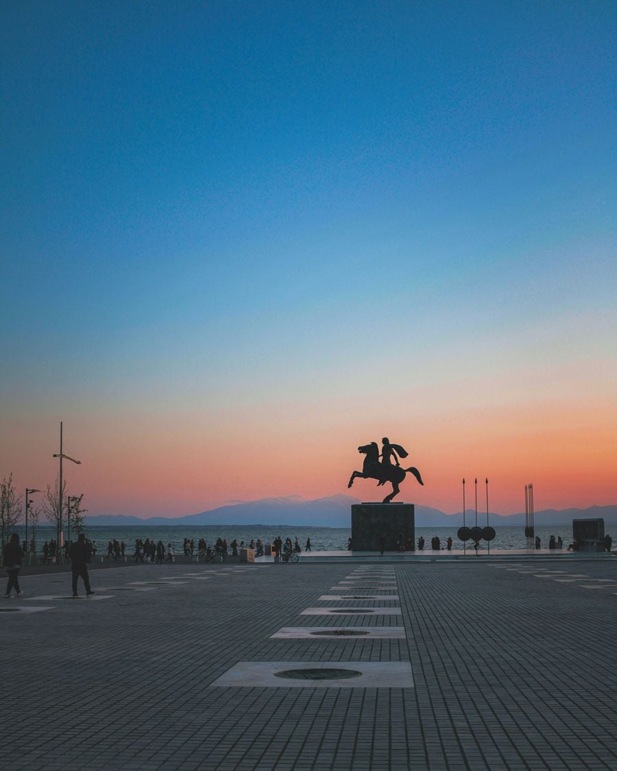 The equestrian statue of Alexander the Great stands silhouetted against a vivid sunset on Thessaloniki’s waterfront. A vast paved plaza leads towards the monument, where pedestrians gather beside the Thermaic Gulf, while distant mountains form a hazy horizon beneath a sky transitioning from deep blue to soft pink and orange.