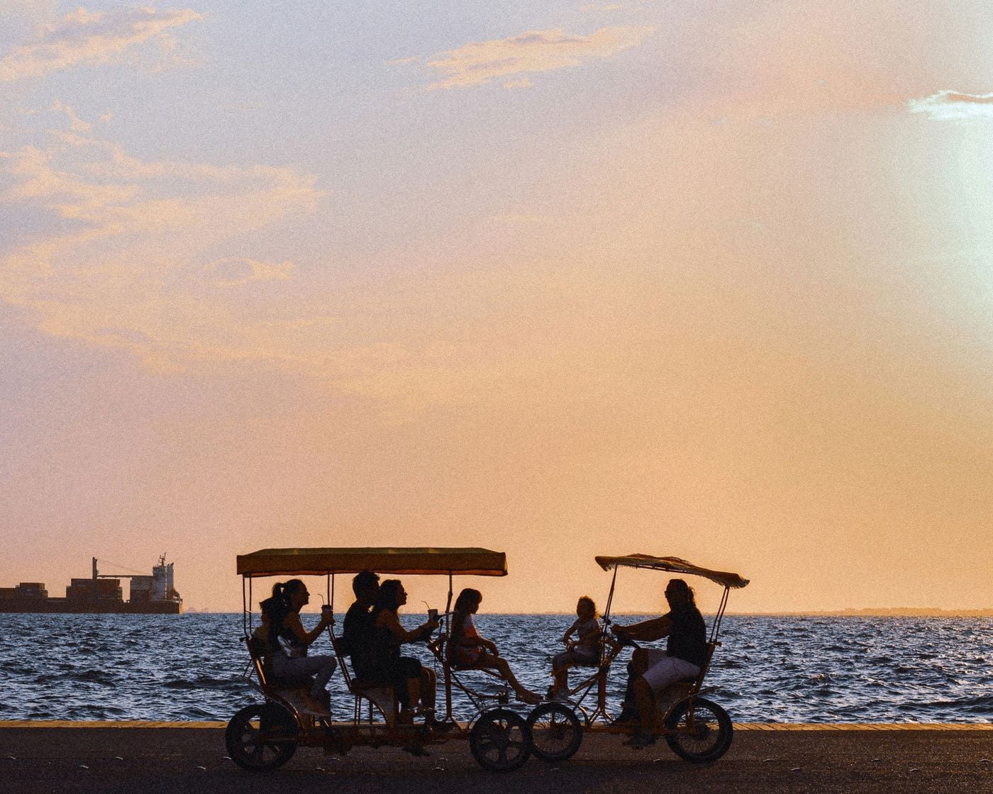 Silhouetted figures ride four-wheeled pedal carts along Thessaloniki’s waterfront promenade at sunset, with the Thermaic Gulf stretching behind them. The warm orange-pink sky, dark sea and distant industrial structures create a nostalgic, cinematic composition capturing the city’s leisurely seaside rhythm.