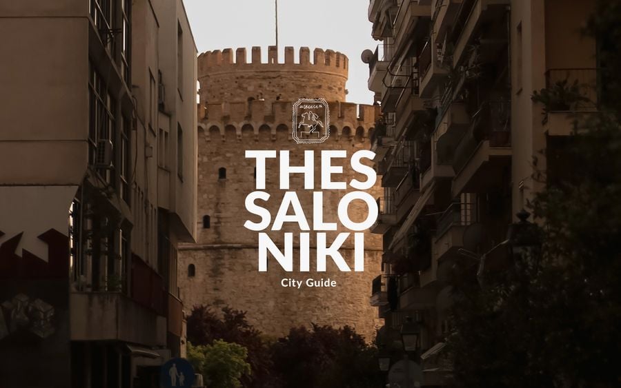 Cover image for Yatzer’s Thessaloniki City Guide, featuring the historic White Tower framed by closely packed urban buildings and shadowed balconies. Warm late-afternoon light illuminates the medieval stone landmark, while bold white typography spelling “THESSALONIKI City Guide” overlays the photograph.