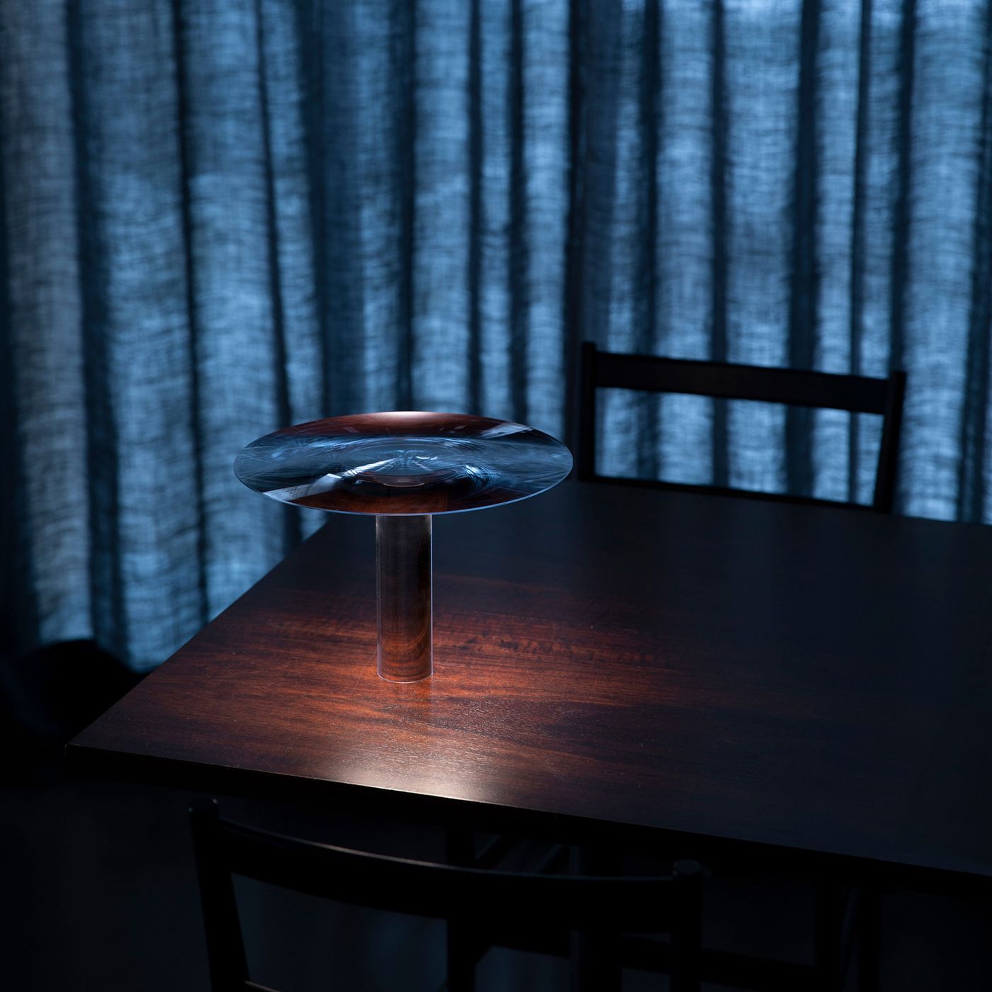 A sculptural table lamp with a lens-like glass top and slender cylindrical base, casting warm light onto a wooden table against a softly draped curtain backdrop A sculptural table lamp with a lens-like glass top and slender cylindrical base, casting warm light onto a wooden table against a softly draped curtain backdrop