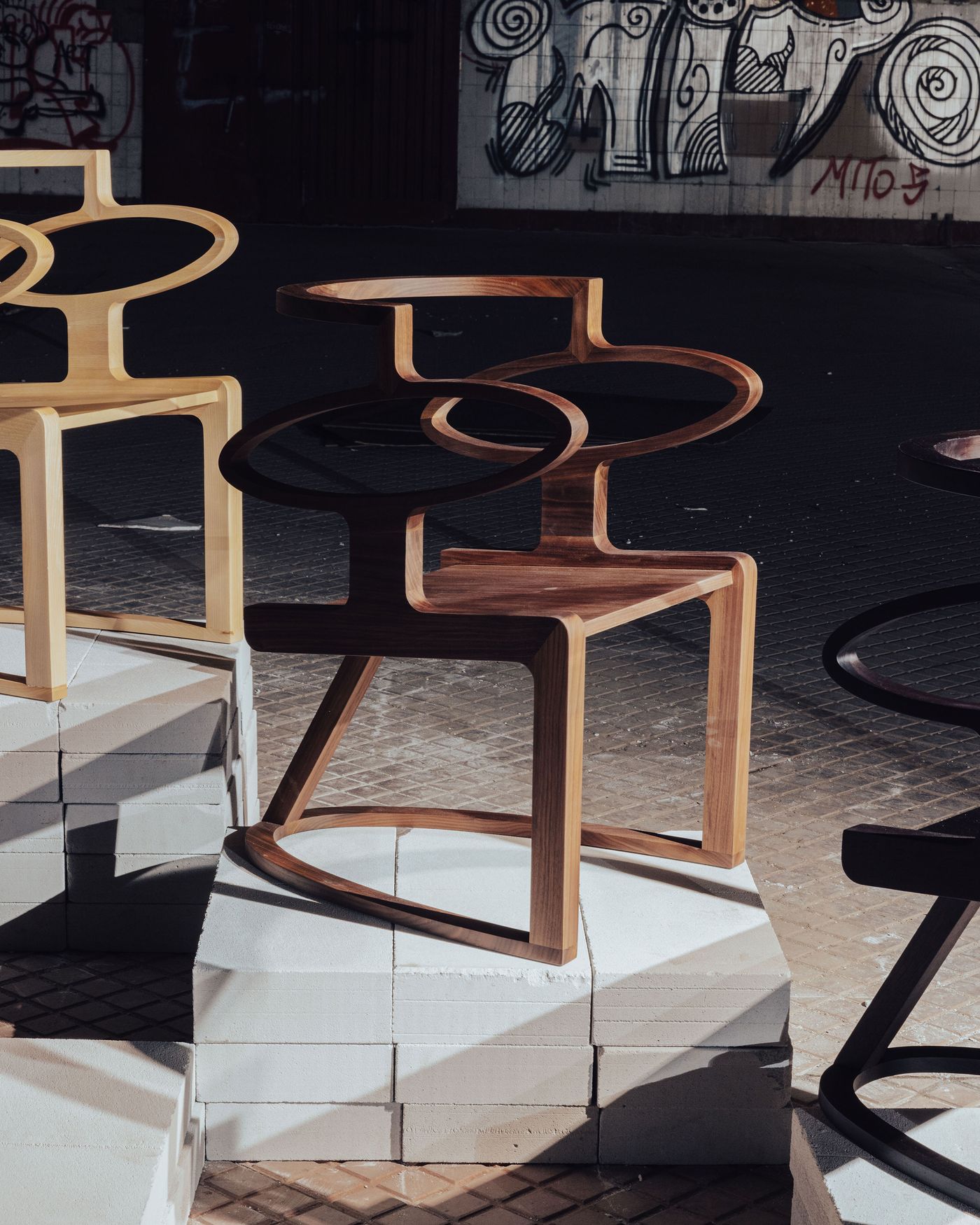 Organic wooden chairs by Abreham, with bold sculptural curves and negative space, glow under the sunlight against a gritty, urban backdrop. Organic wooden chairs by Abreham, with bold sculptural curves and negative space, glow under the sunlight against a gritty, urban backdrop.
