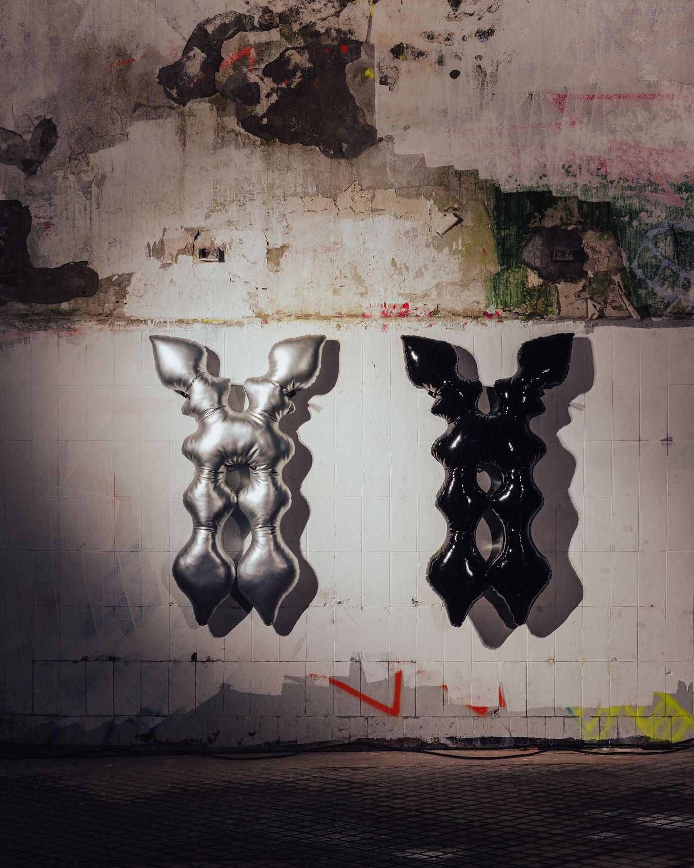 Two sculptural cushions by Claire Duport, one in metallic silver and one in glossy black, hang against a raw, graffiti-marked wall, blending softness with urban decay. Two sculptural cushions by Claire Duport, one in metallic silver and one in glossy black, hang against a raw, graffiti-marked wall, blending softness with urban decay.