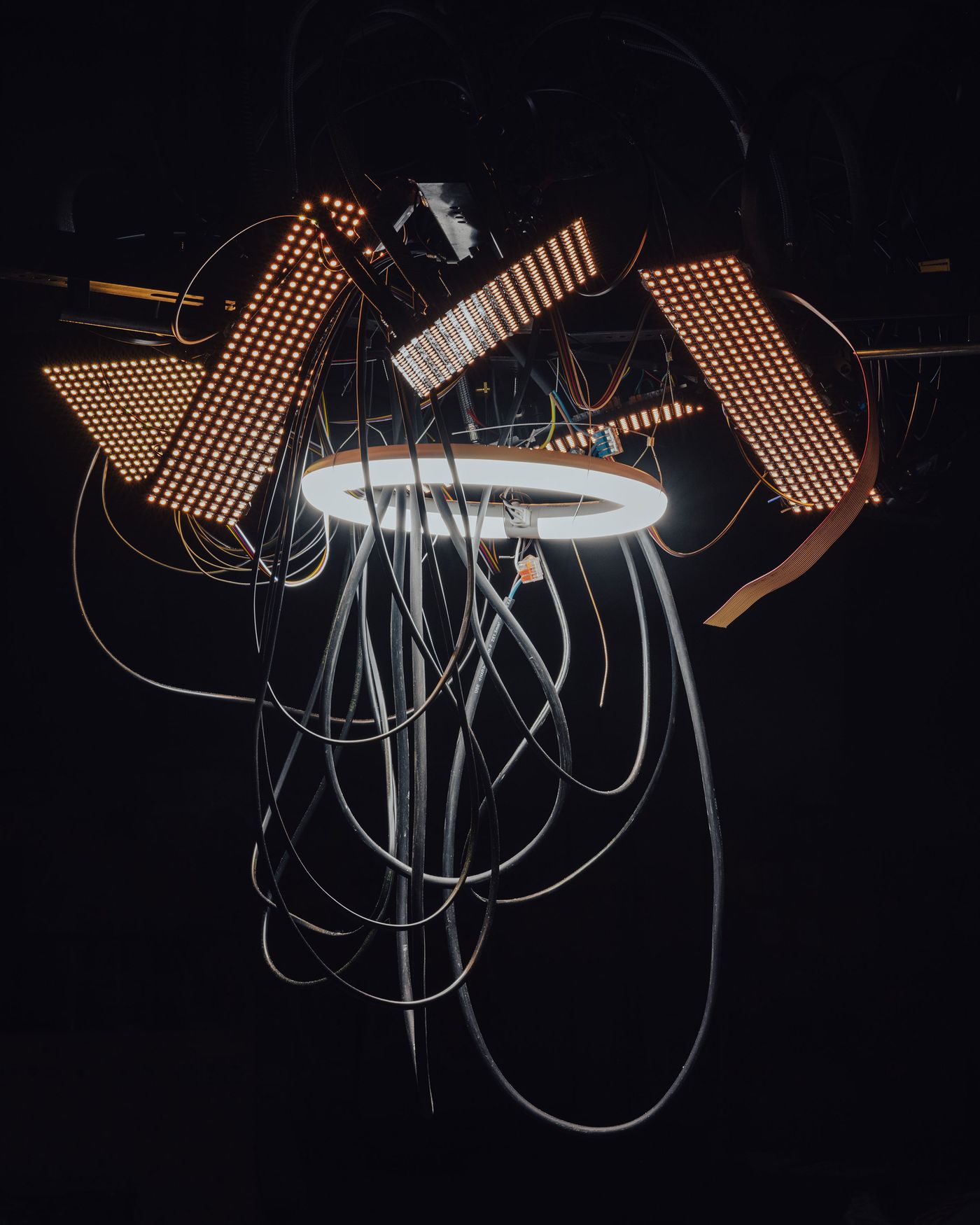 A chaotic tangle of glowing cables and LED panels forms a futuristic, sculptural chandelier suspended in darkness. A chaotic tangle of glowing cables and LED panels forms a futuristic, sculptural chandelier suspended in darkness.