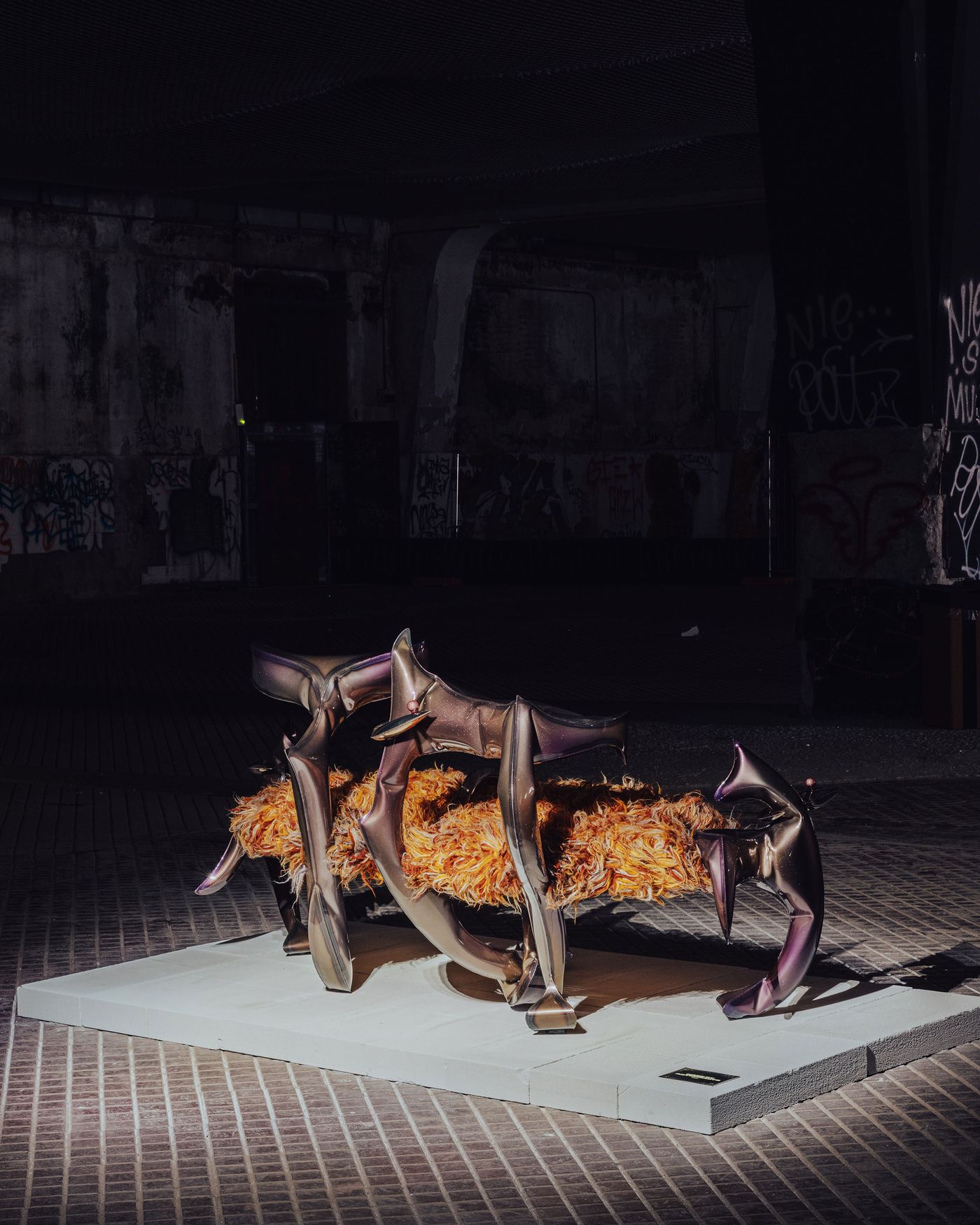 A sculptural bench with twisted, polished steel legs and a vibrant orange flokati wool seat, dramatically staged in a dark, abandoned, graffiti-covered industrial space. A sculptural bench with twisted, polished steel legs and a vibrant orange flokati wool seat, dramatically staged in a dark, abandoned, graffiti-covered industrial space.