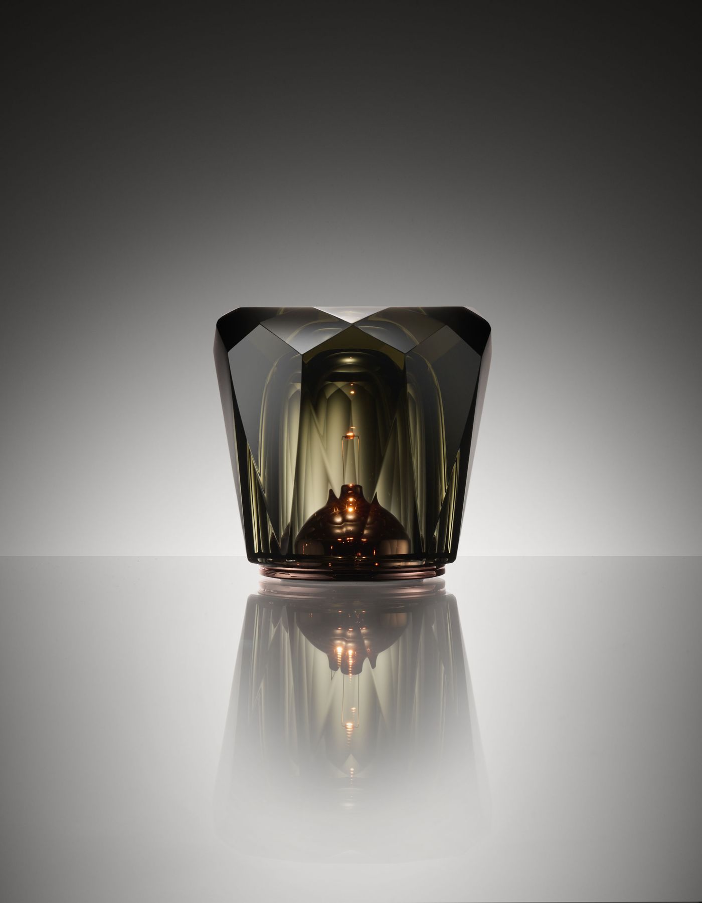 A faceted, smoky glass table lamp with a jewel-like geometric design sits on a reflective surface, softly glowing from within and casting intricate reflections and highlights against a gradient gray background. A faceted, smoky glass table lamp with a jewel-like geometric design sits on a reflective surface, softly glowing from within and casting intricate reflections and highlights against a gradient gray background.