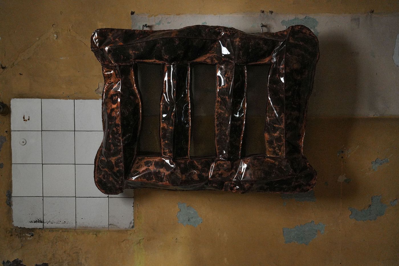 Abstract wall-mounted sculpture with an organic, inflated form and glossy, mottled brown-black texture, displayed against a distressed yellow wall with white tiles.