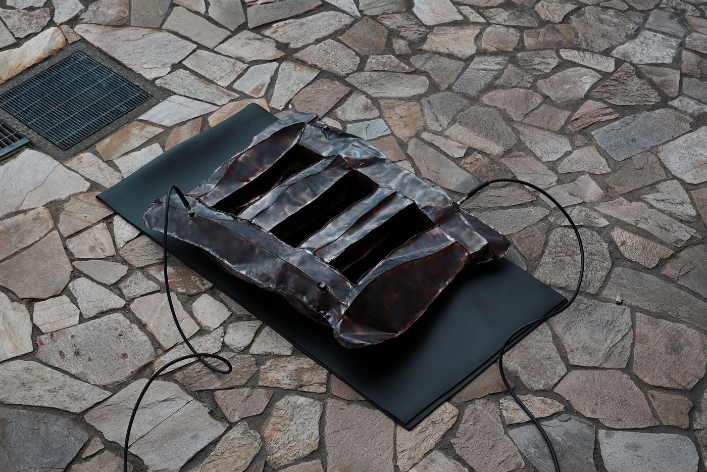 A hammered, bronze-toned metal sculpture with hollow sections placed on a black mat over a stone floor, connected to black wires.