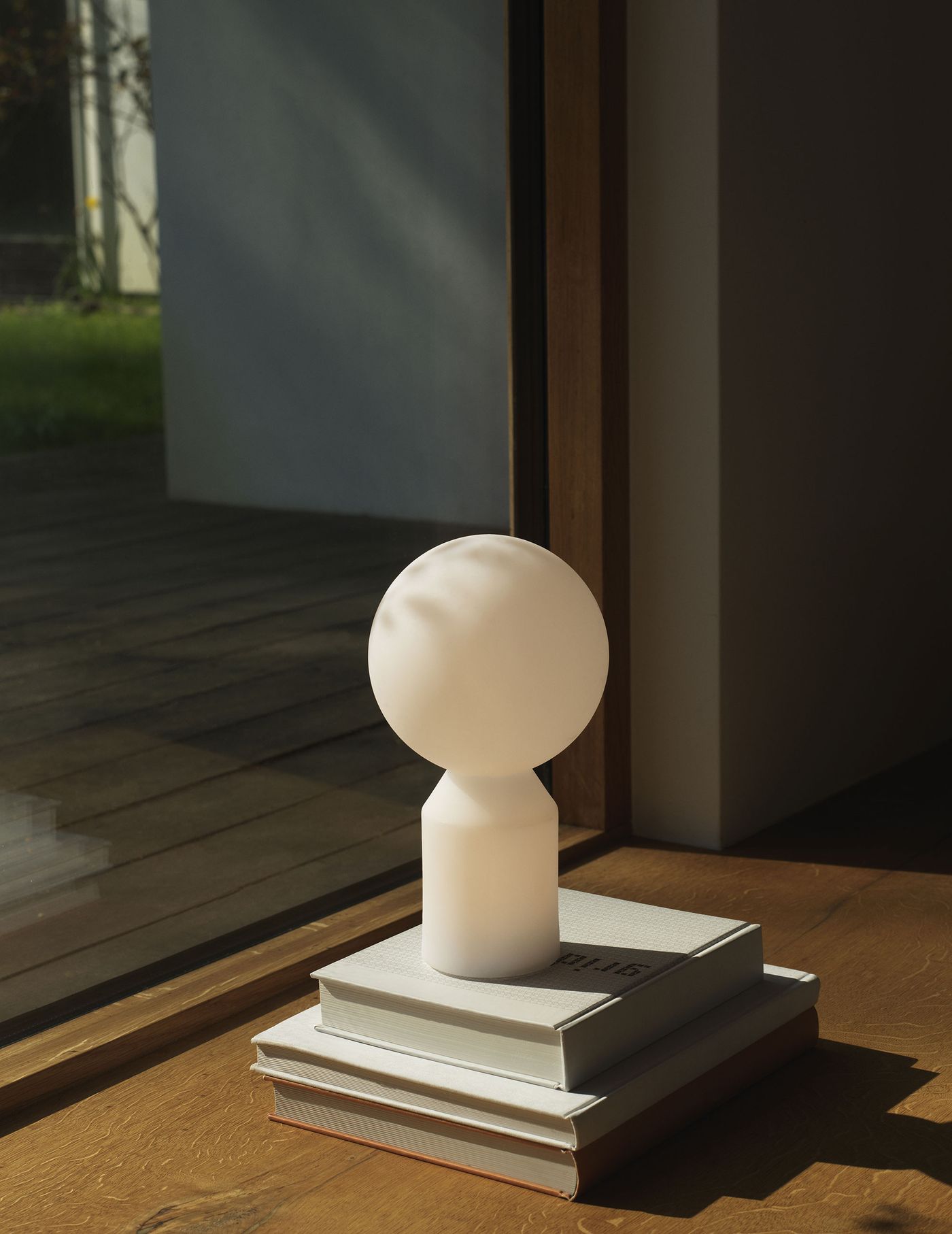 A sculptural, frosted glass lamp with a rounded top and faceted base sits atop a stack of books by a sunlit window, blending minimalism with quiet character.