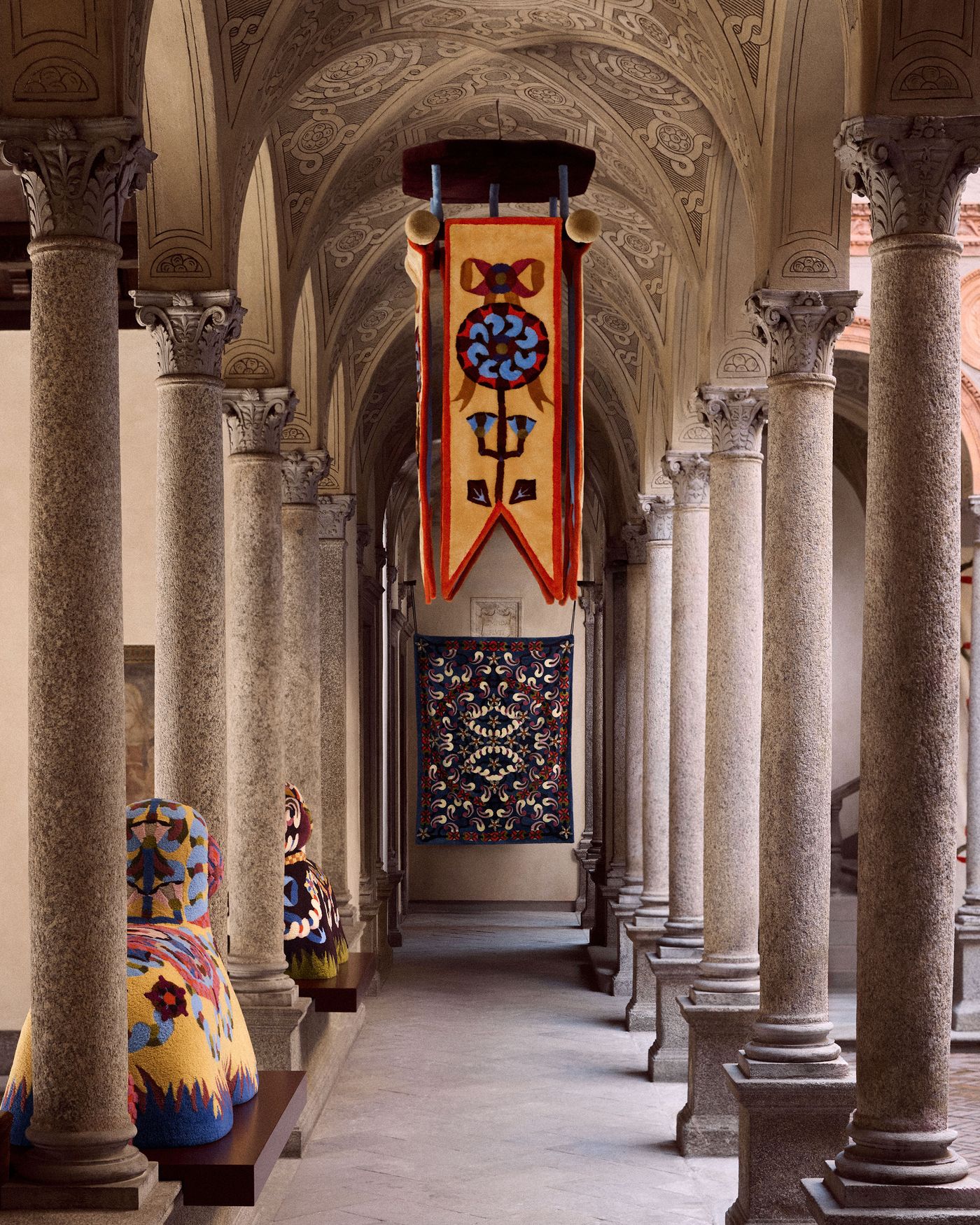 A grand cloister with ornate columns and vaulted ceilings displays colorful embroidered banners and abstract, textile animal sculptures. A grand cloister with ornate columns and vaulted ceilings displays colorful embroidered banners and abstract, textile animal sculptures.