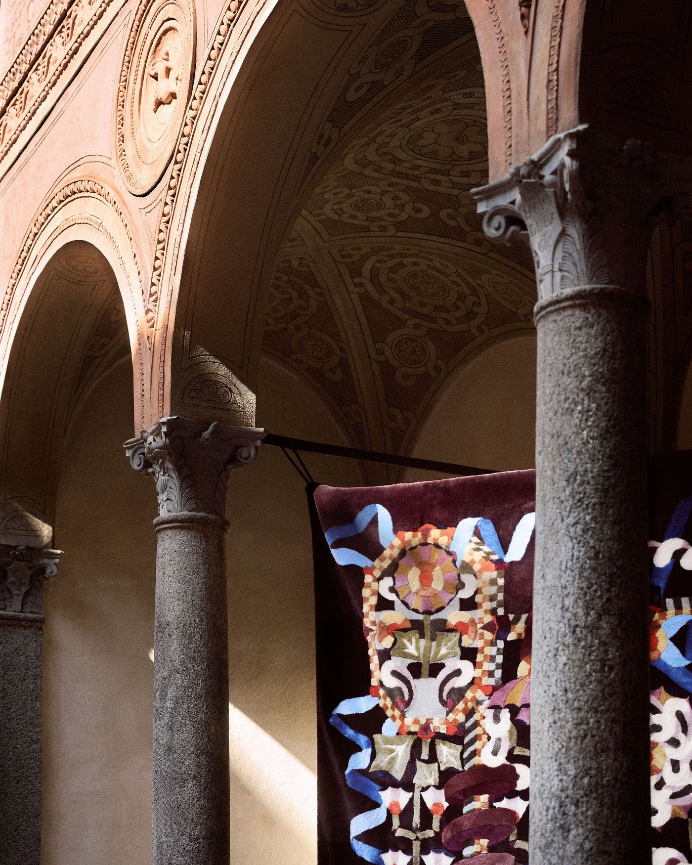 Close-up of a richly embroidered tapestry hanging under intricately carved stone arches, bathed in soft natural light.
