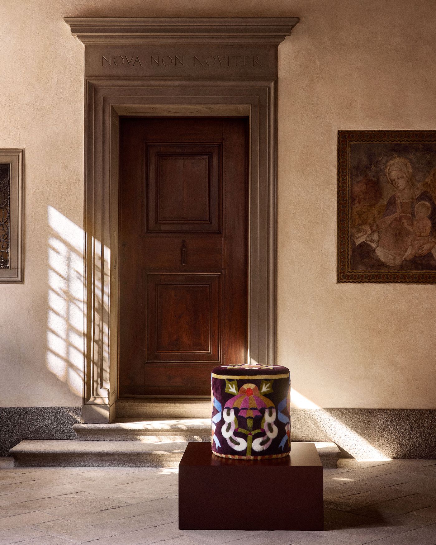 A velvet-upholstered cylindrical stool with vivid, abstract embroidery sits on a raised platform by a traditional wooden door.