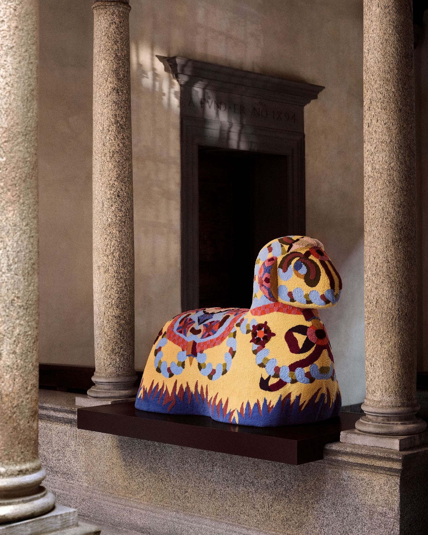 A vibrant, yellow textile animal sculpture with playful embroidery details stands between classic stone columns.