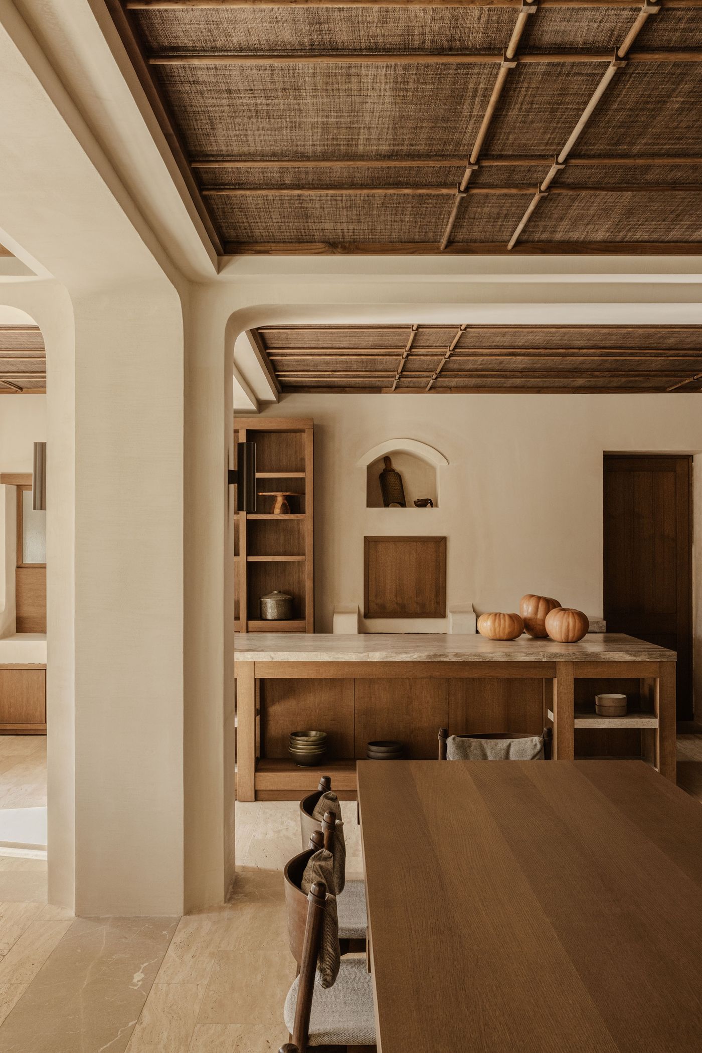 A serene view of a minimalist kitchen featuring natural textures, travertine floors, and wooden accents, with pumpkins arranged on a central island.