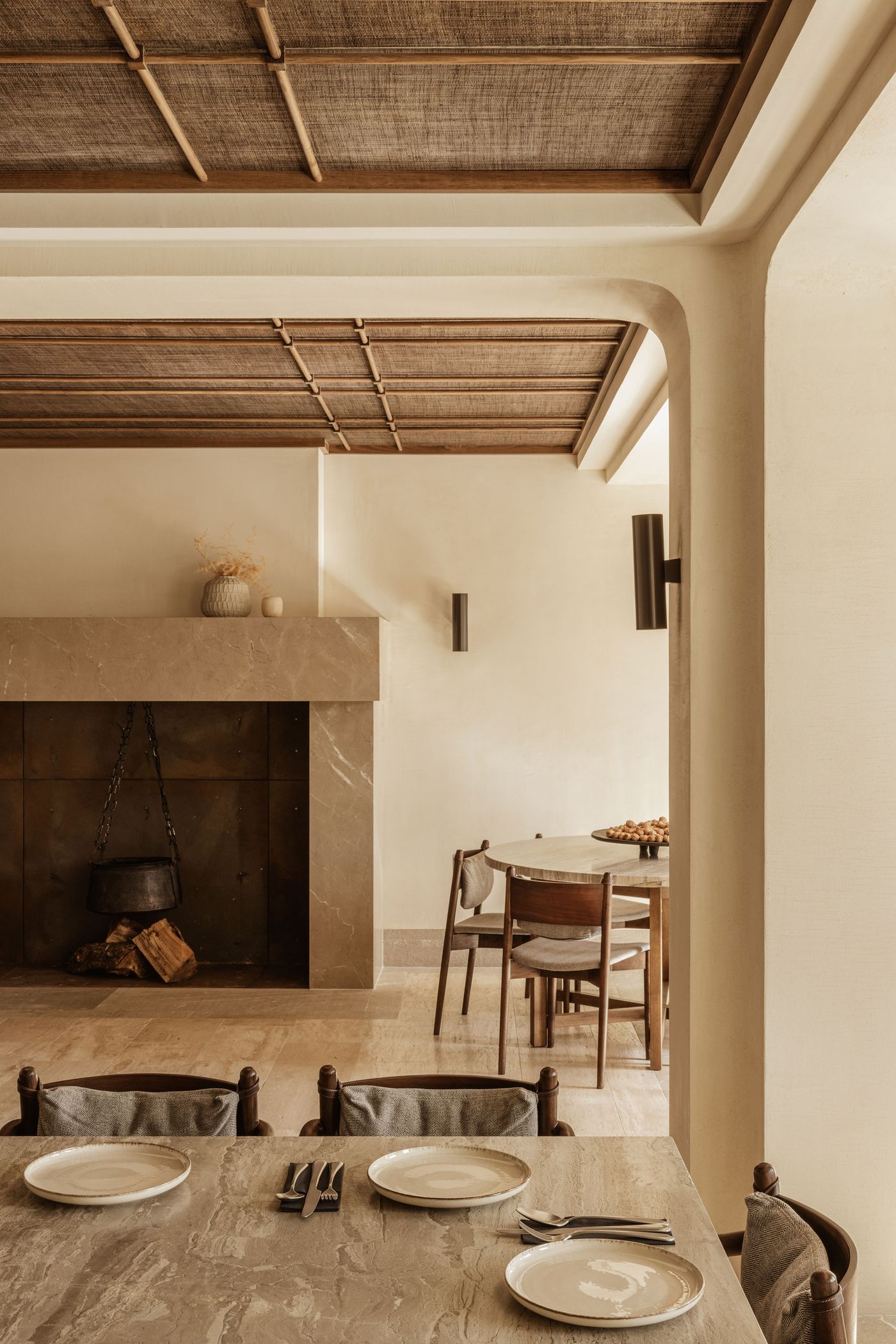 Earth-toned dining space with a stone fireplace, round marble table, and artisanal ceramic decor under a woven ceiling.