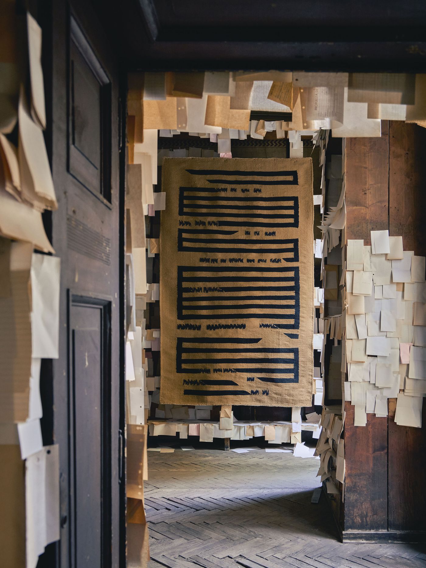A cozy corridor wrapped in floating sheets of paper leads to a beige woven rug with bold black geometric lines, suspended like a tapestry.