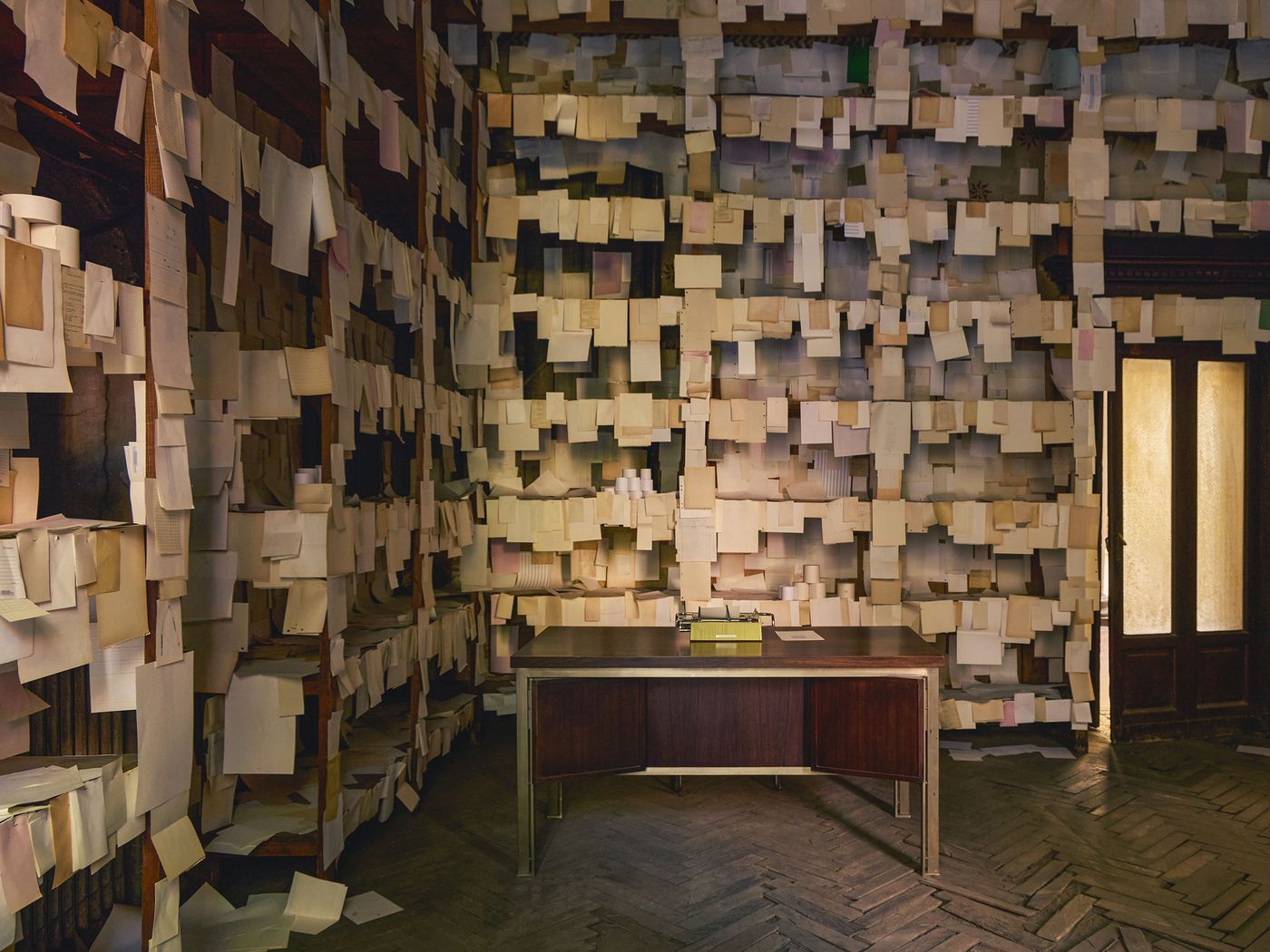 A moody, nostalgic room entirely covered in pinned papers, with a vintage wooden desk and a bright yellow typewriter placed at its center, under soft golden light.