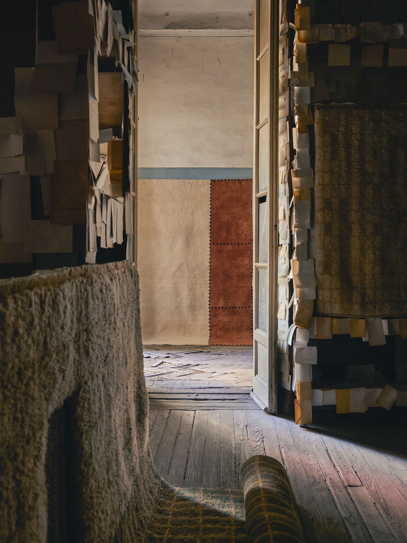 A sun-drenched doorway reveals a minimal room with a two-tone rug in warm rust and cream shades, framed by walls layered with pinned paper scraps.