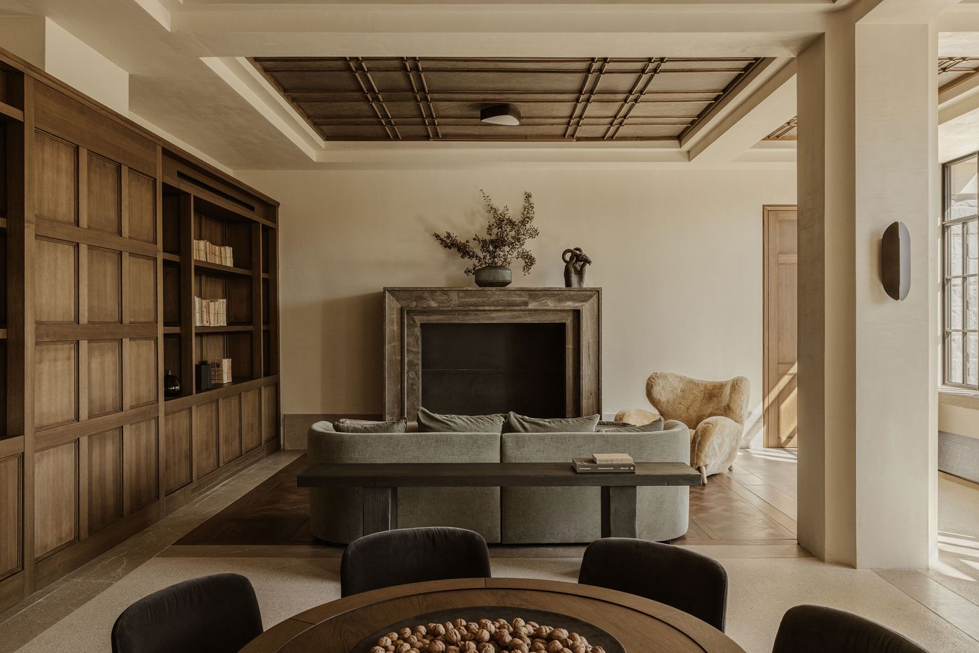 Elegant living room with a muted green velvet sofa, sculptural armchair, and a large fireplace framed by built-in wooden bookshelves.