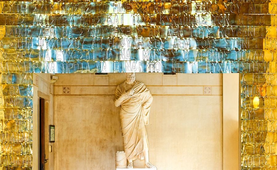 A head-on view of Gabriel Leger's sculptural installation at Villa Kerylos. The cascade of golden metallic sheets, etched with Greek script, creates a translucent doorway. Beyond, a classical statue stands in a sun-drenched corridor with intricate wall friezes and a subtle floor mosaic.