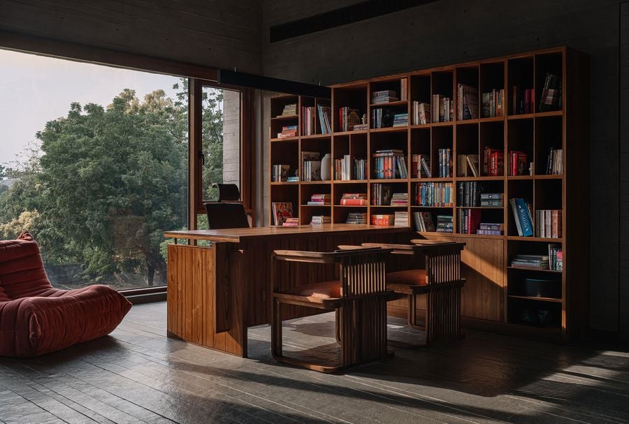 An expansive and inviting study within the MS House, featuring a substantial wooden desk and matching chairs, complemented by a large built-in bookshelf filled with books. Floor-to-ceiling windows provide abundant natural light and sweeping views of lush trees, creating an inspiring and serene work environment. A vibrant red lounge chair adds a playful touch.