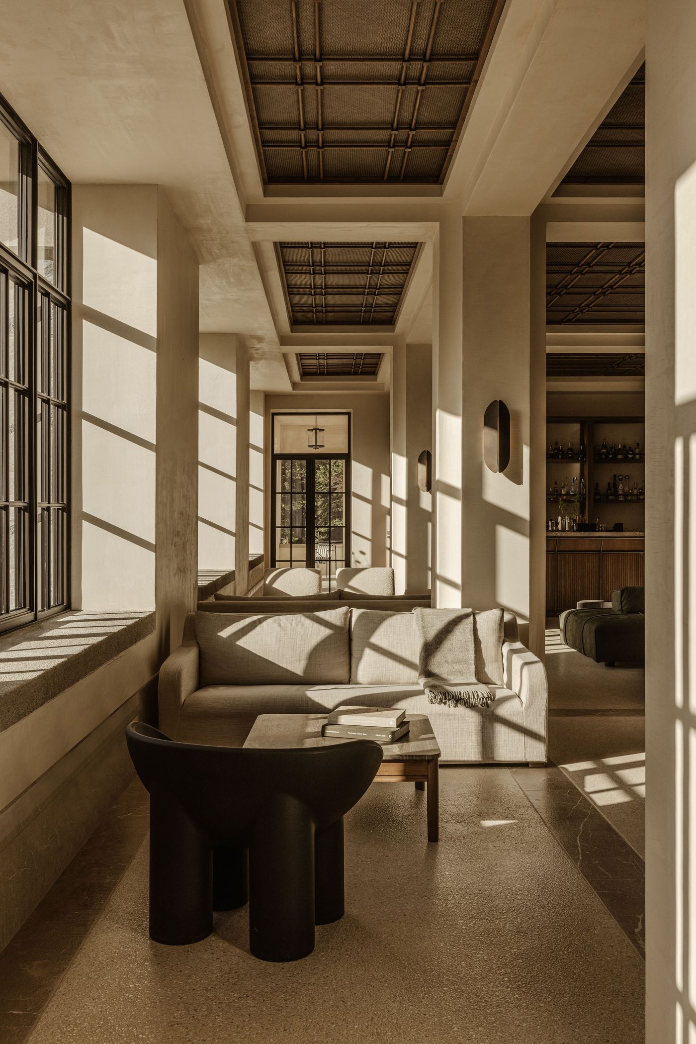 Sunlit corridor with cream-toned sofas, sculptural black chairs, and long shadows streaming through grid-framed windows.