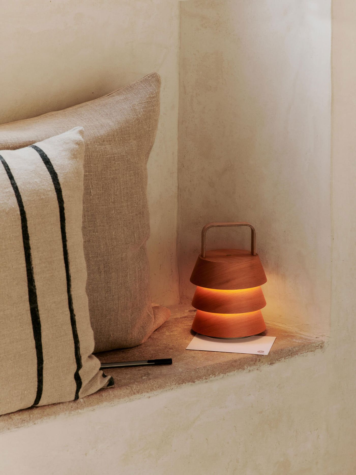 A wooden Luver Portable Lamp by Ferm Living emits a soft, warm glow on a stone ledge beside a neutral-toned linen cushion with black stripes, creating a serene, earthy ambiance.