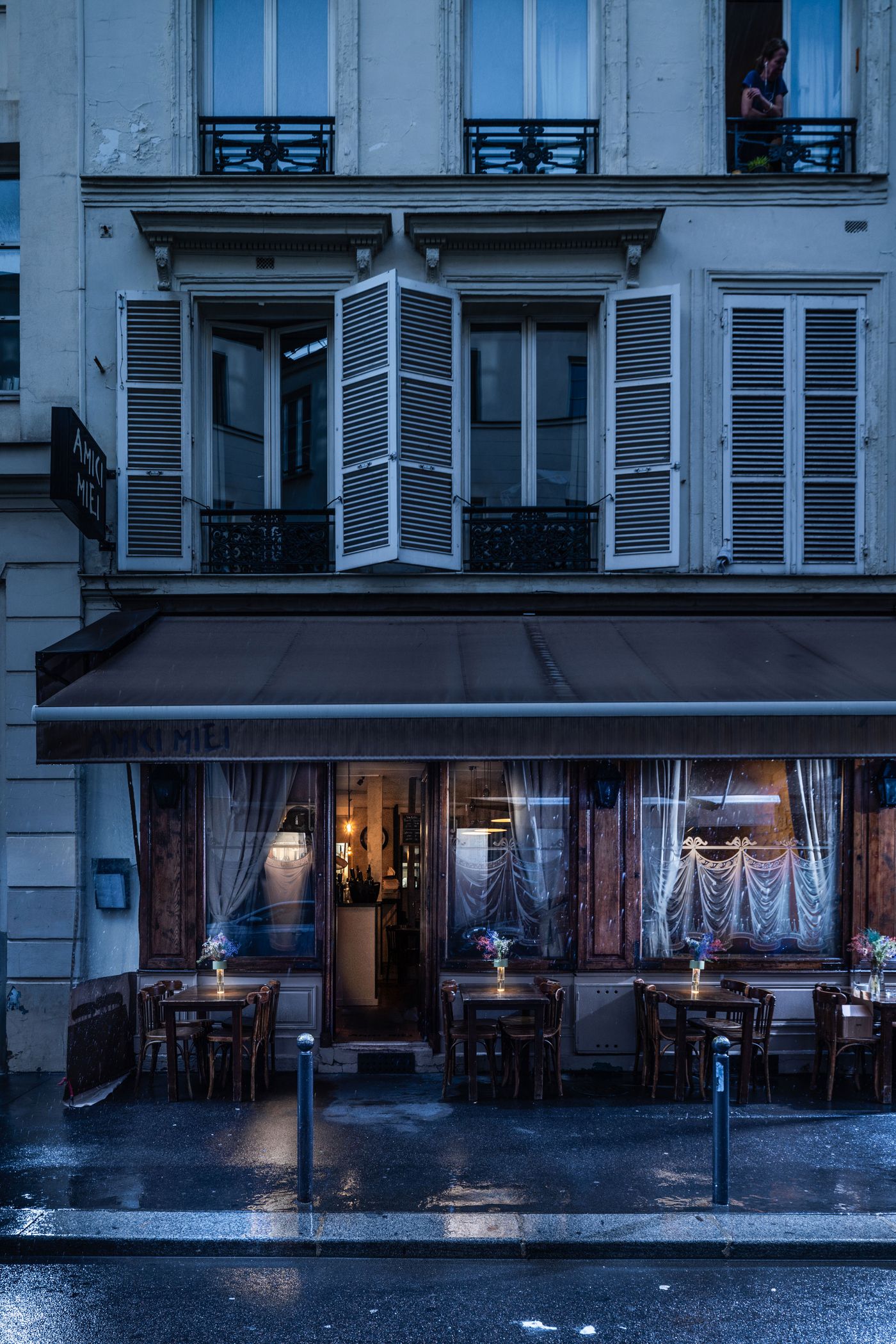 A moody street view of a rain-kissed Parisian café named “Amici Miei” at twilight, with warm light glowing from within and a row of wooden tables adorned with floral arrangements and illuminated Fleur lamps placed on the wet pavement outside. A moody street view of a rain-kissed Parisian café named “Amici Miei” at twilight, with warm light glowing from within and a row of wooden tables adorned with floral arrangements and illuminated Fleur lamps placed on the wet pavement outside.