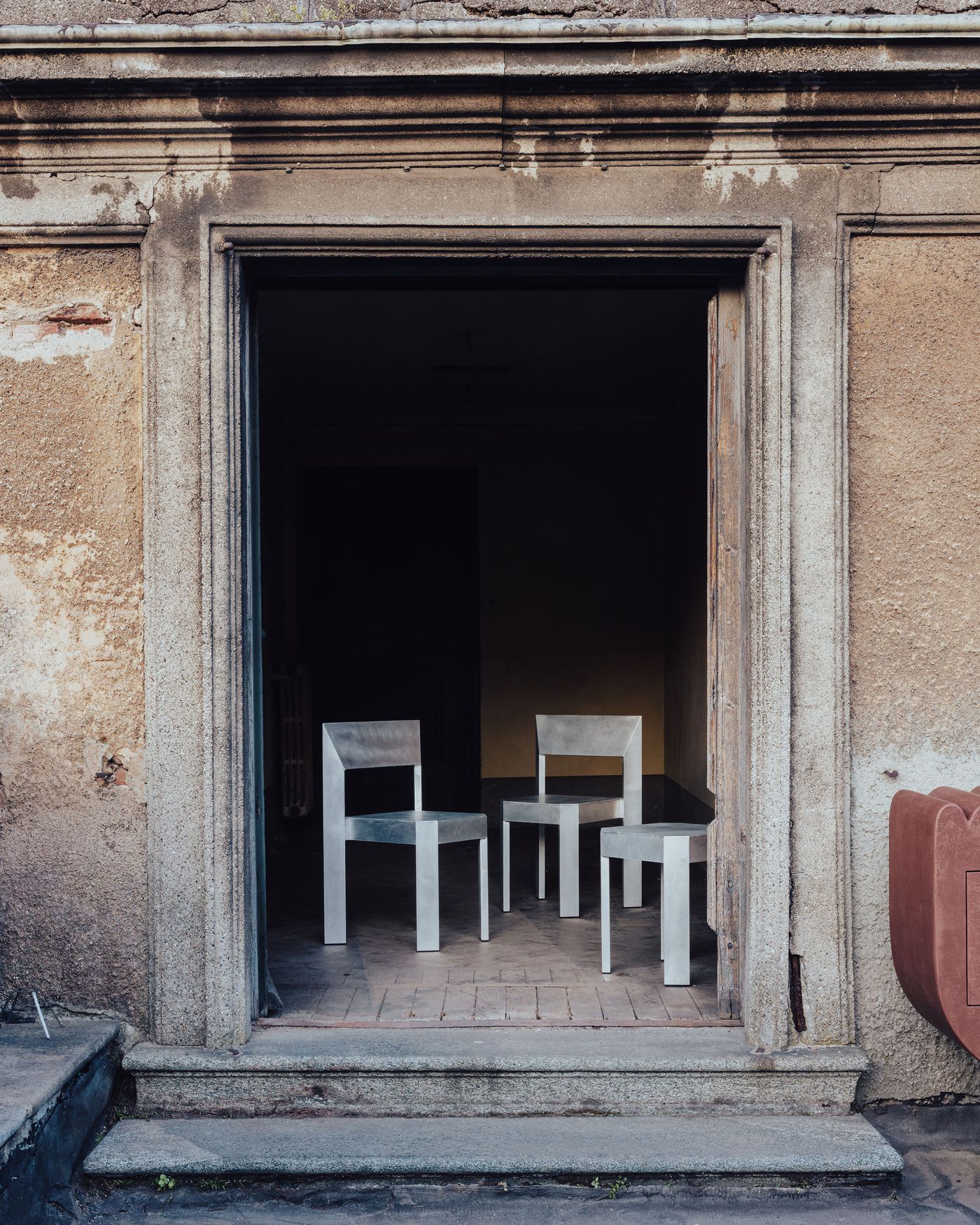 Three minimalist, angular chairs in a raw metallic finish are placed inside a weathered stone doorway of an old, textured building, blending stark modern design with a crumbling historic backdrop. Three minimalist, angular chairs in a raw metallic finish are placed inside a weathered stone doorway of an old, textured building, blending stark modern design with a crumbling historic backdrop.