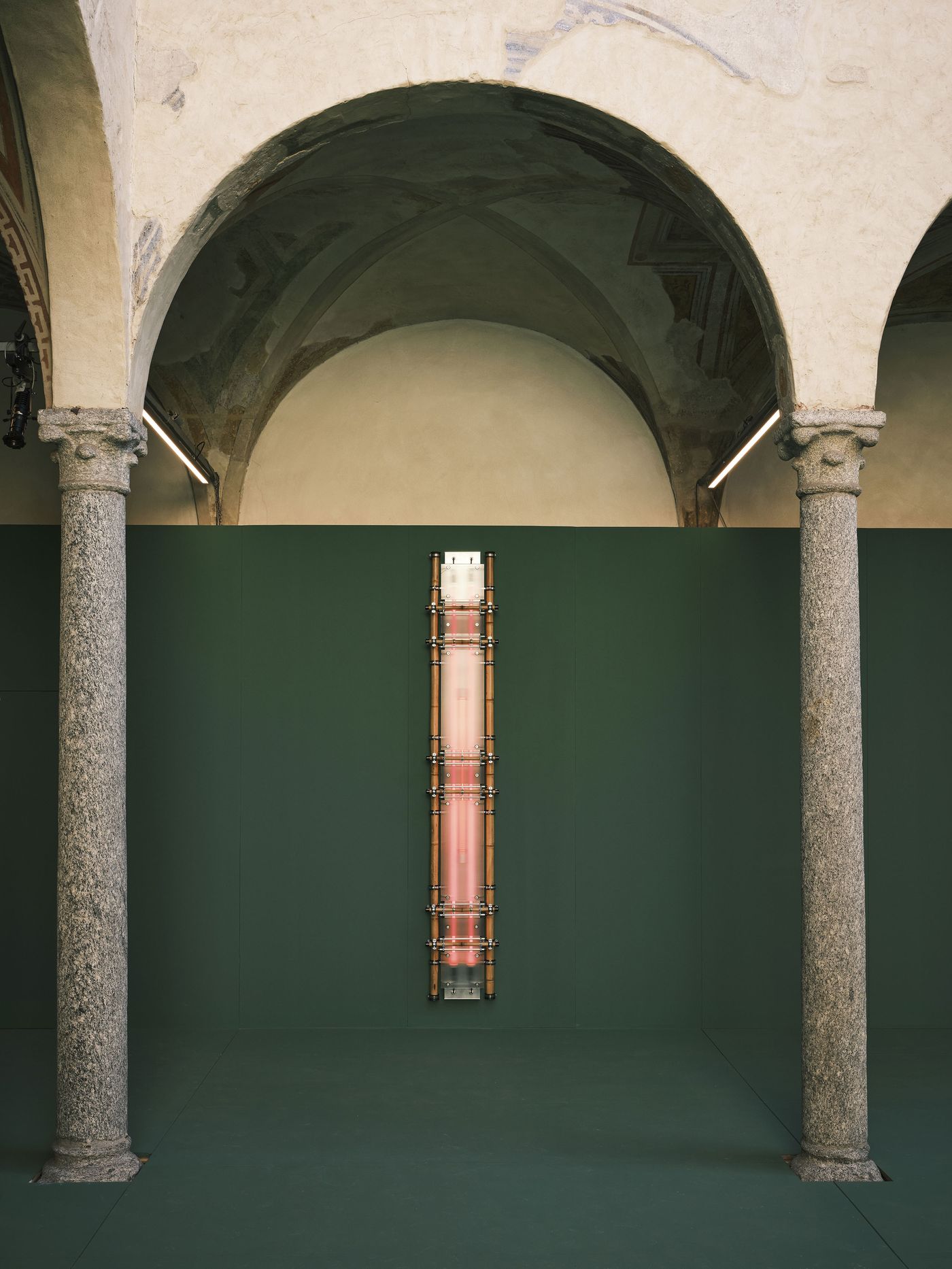 Installation view of a vertically mounted sculptural structure with bamboo rods, metal clamps, and a translucent glowing panel, centered on a dark green wall beneath arched stone architecture. Installation view of a vertically mounted sculptural structure with bamboo rods, metal clamps, and a translucent glowing panel, centered on a dark green wall beneath arched stone architecture.