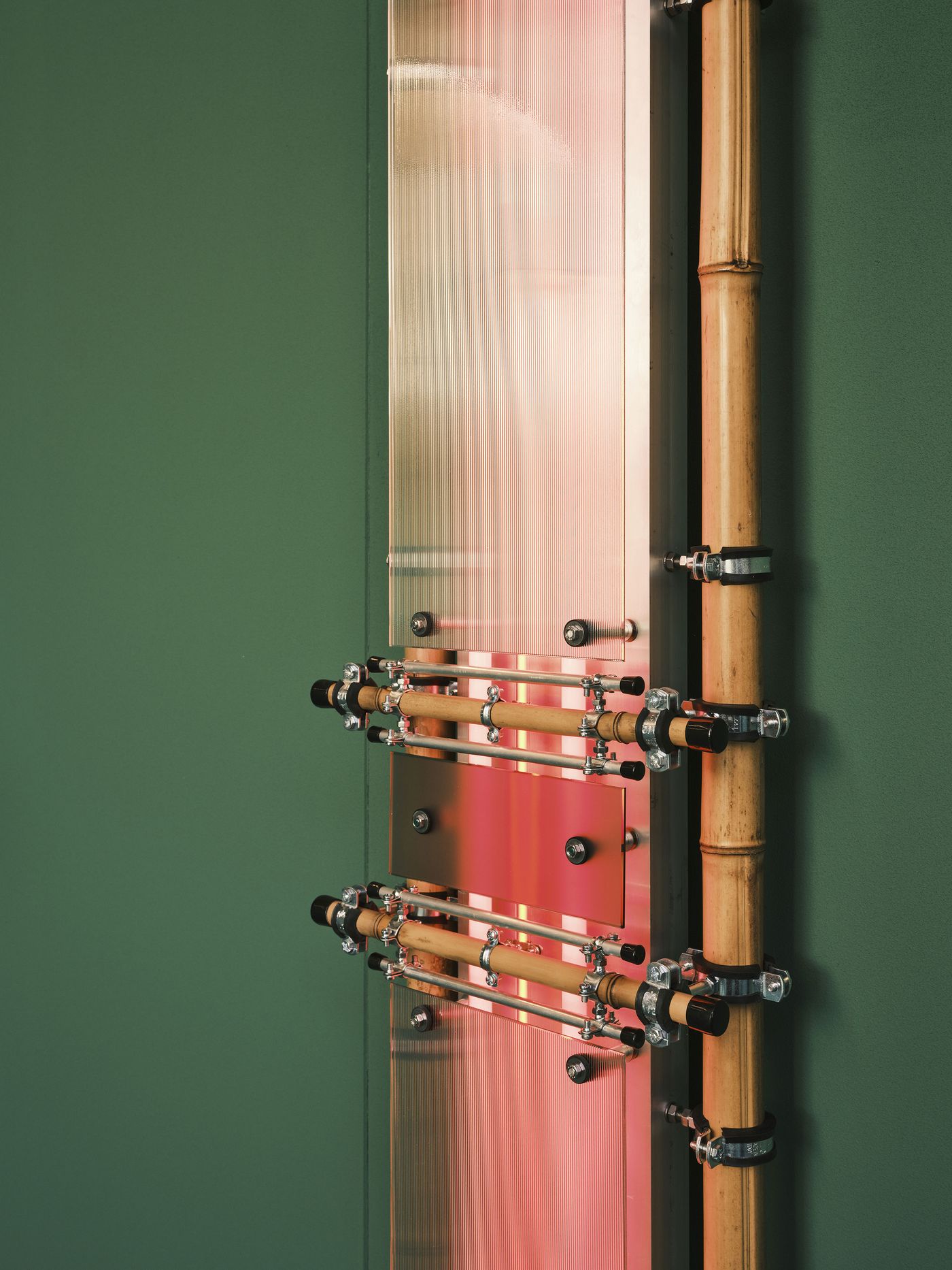 Close-up of a light installation combining natural bamboo, industrial metal fixtures, and a glowing iridescent surface, creating a contrast of organic and engineered aesthetics against a green background. Close-up of a light installation combining natural bamboo, industrial metal fixtures, and a glowing iridescent surface, creating a contrast of organic and engineered aesthetics against a green background.