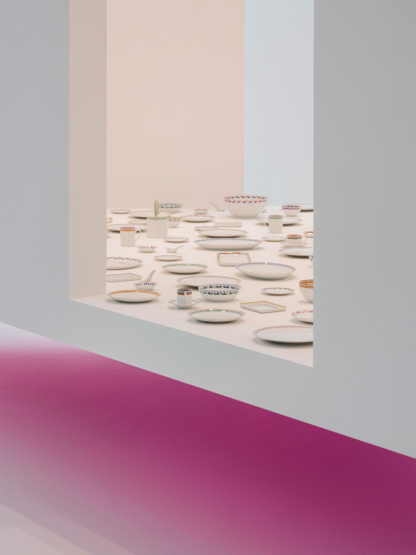 A floating white display cube reveals a meticulously arranged collection of Hermès tableware—plates, bowls, and cups adorned with colorful graphic patterns—set against soft pastel walls and a gradient magenta floor that adds a dreamy, gallery-like ambience. A floating white display cube reveals a meticulously arranged collection of Hermès tableware—plates, bowls, and cups adorned with colorful graphic patterns—set against soft pastel walls and a gradient magenta floor that adds a dreamy, gallery-like ambience.