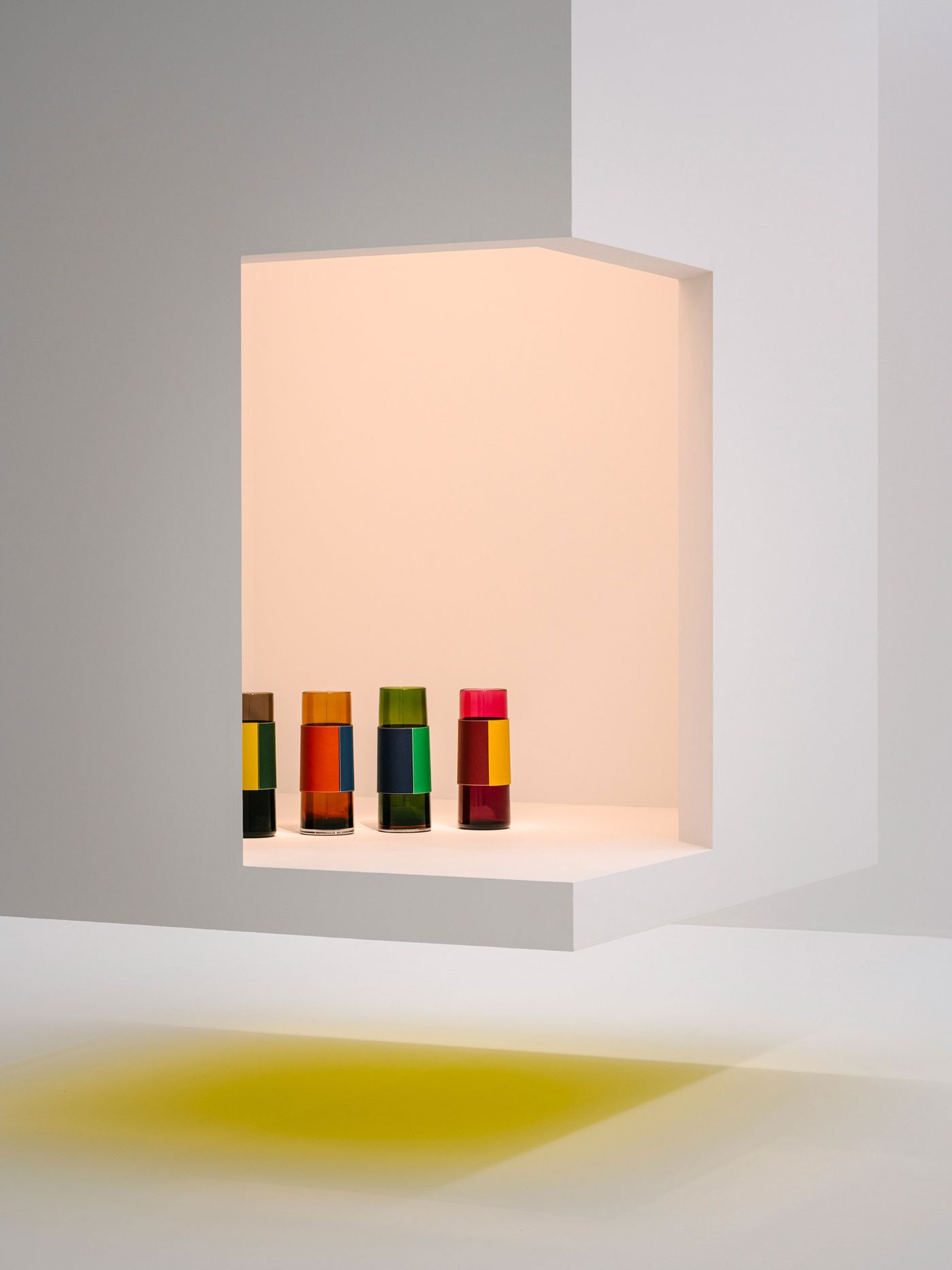 A minimalist cut-out display niche showcases a row of vibrant, color-blocked Hermès glass tumblers in bold jewel tones, casting a soft yellow glow on the white floor below—pure geometry meets playful luxury. A minimalist cut-out display niche showcases a row of vibrant, color-blocked Hermès glass tumblers in bold jewel tones, casting a soft yellow glow on the white floor below—pure geometry meets playful luxury.