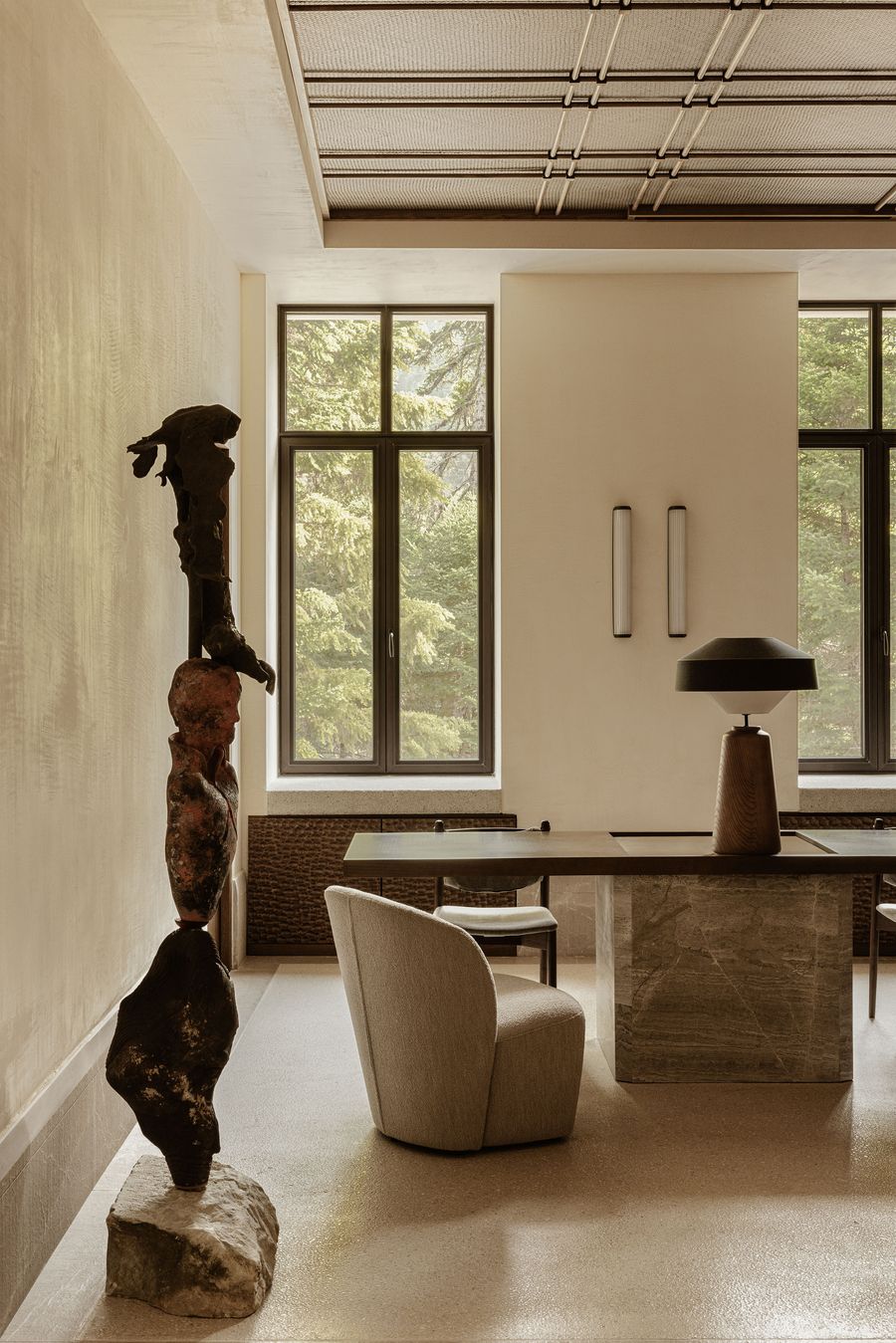 Interior scene with a sculptural totem-like artwork beside a desk setup, flanked by tall windows that reveal a dense pine forest beyond. Interior scene with a sculptural totem-like artwork beside a desk setup, flanked by tall windows that reveal a dense pine forest beyond.