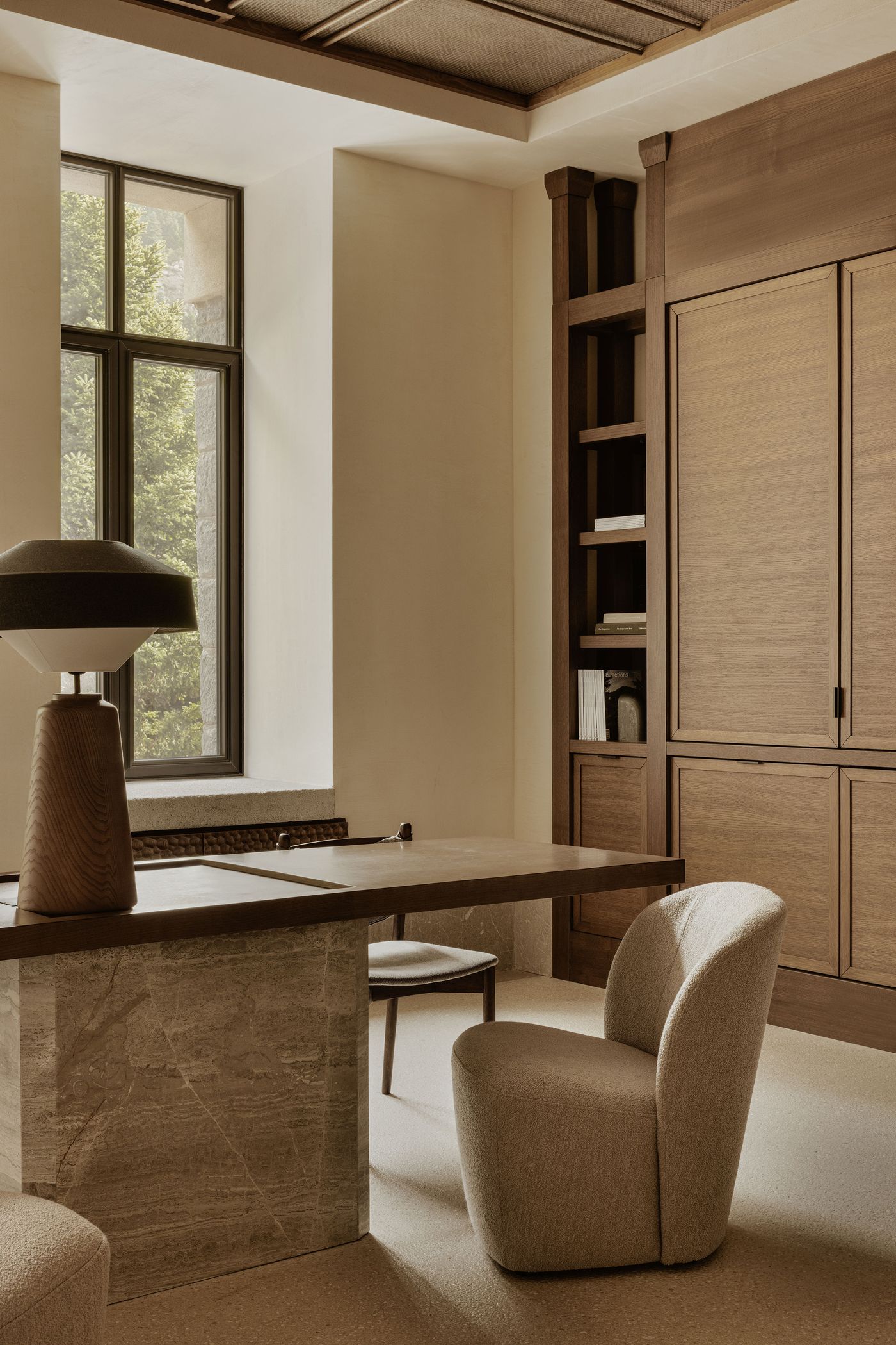 The Manna hotel's reception featuring a travertine desk, boucle armchair, and sculptural wooden lamp set against built-in oak cabinetry and a window framing lush greenery.