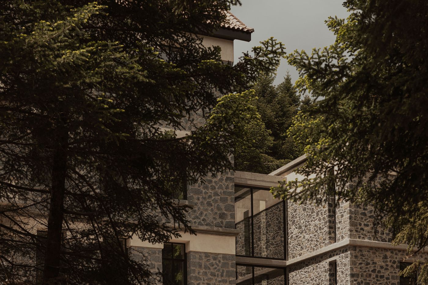 Close-up of the Manna Arcadia hotel’s stone facade partially concealed by fir trees, showcasing a minimalist extension in glass and steel that blends into the mountainous landscape. Close-up of the Manna Arcadia hotel’s stone facade partially concealed by fir trees, showcasing a minimalist extension in glass and steel that blends into the mountainous landscape.