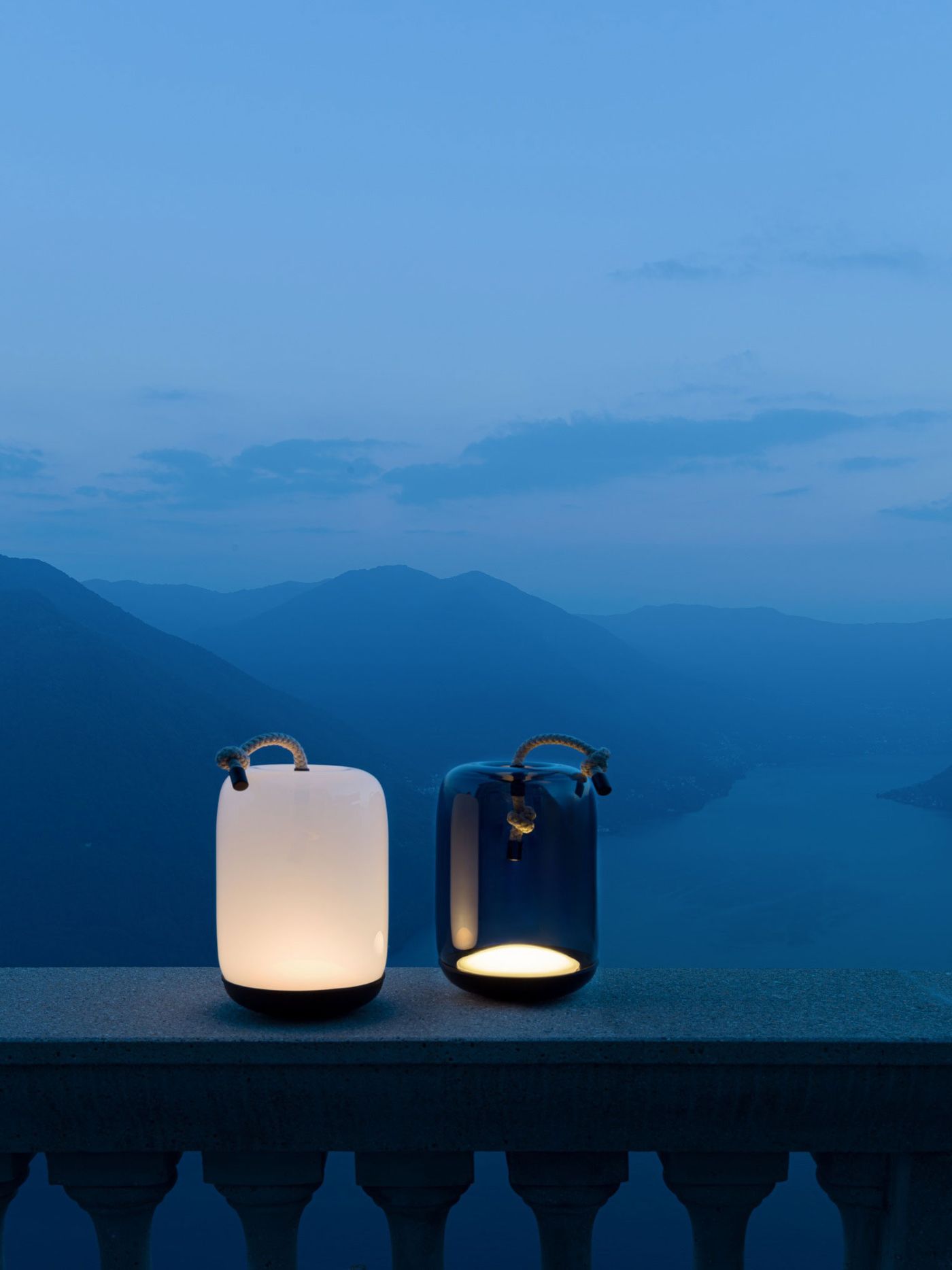 Two sculptural glass lanterns by Brokis glowing softly at twilight, perched above a dreamy mountain lake panorama. Two sculptural glass lanterns by Brokis glowing softly at twilight, perched above a dreamy mountain lake panorama.
