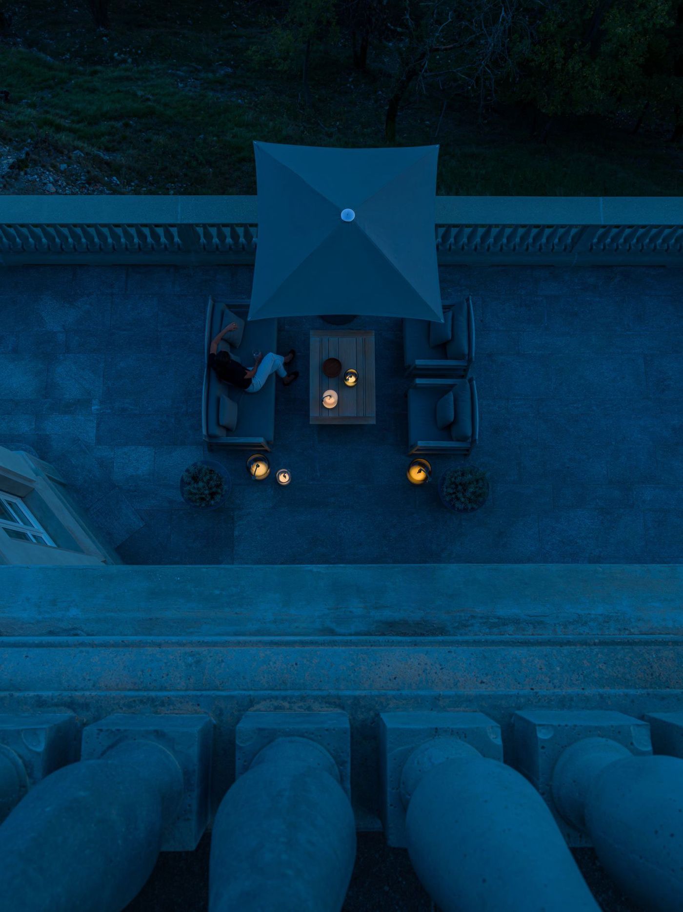 A twilight terrace scene viewed from above, where several glowing Knot Battery lamps by Brokis gently illuminate a lounge setup beneath a square umbrella. A twilight terrace scene viewed from above, where several glowing Knot Battery lamps by Brokis gently illuminate a lounge setup beneath a square umbrella.