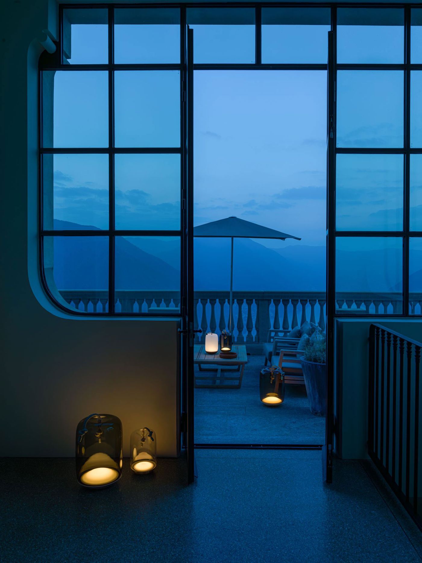 An atmospheric view through large glass doors at dusk, revealing a mountain terrace adorned with multiple Knot Battery lamps in various sizes, casting a soft golden glow. An atmospheric view through large glass doors at dusk, revealing a mountain terrace adorned with multiple Knot Battery lamps in various sizes, casting a soft golden glow.