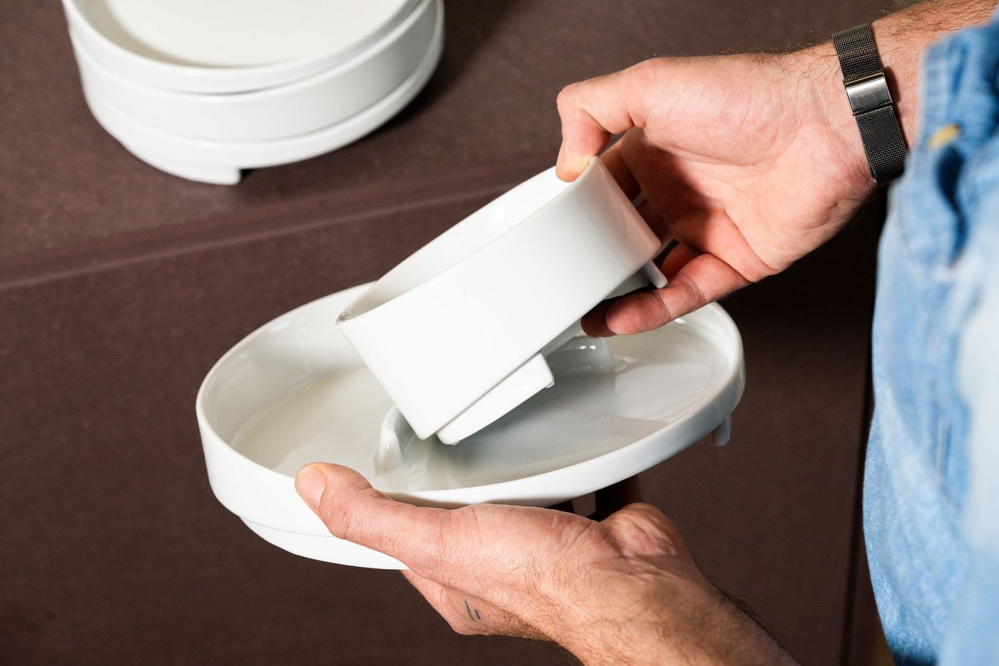 Hands demonstrating the modular functionality of the white ceramic tableware by lifting one bowl from another.